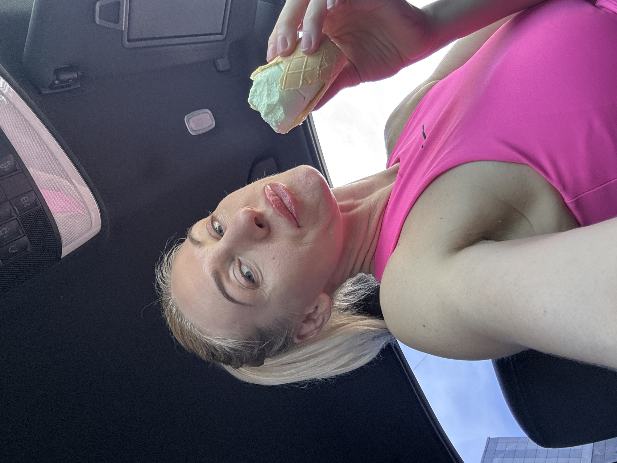 A fair-skinned blonde woman wearing a pink top is holding a green-tinted ice cream cone in a car. The interior of the car has a black ceiling and the car's visor and controls are visible. The woman is looking at the camera. A building is visible through the car's window. The location is Unknown, Unknown.