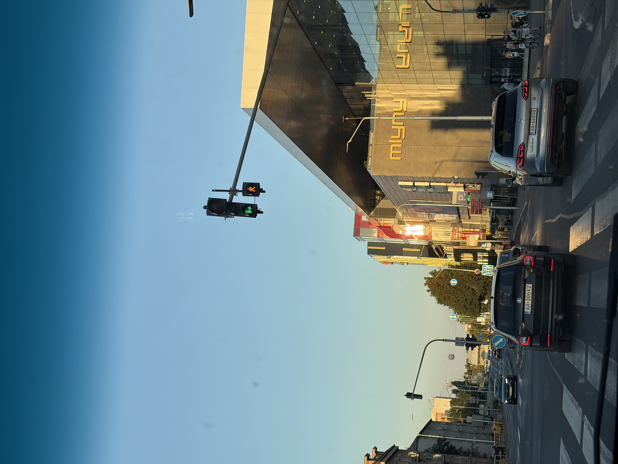 The media file depicts a street scene in Nitra, Slovakia, during what appears to be late afternoon or early evening. The sun casts long shadows, indicating a low angle of light. The sky is a clear, pale blue, suggesting fair weather.
The scene is dominated by a modern, angular building with a reflective facade, displaying yellow text that reads "Mlynar" and "terasa". In front of this building, traffic lights are visible, with one showing a green pedestrian signal and another a yellow pedestrian signal. Several vehicles are on the road, including a silver Volkswagen Touareg with a license plate "AA*648VD" and a dark-colored Skoda Octavia with a license plate "AA 306 KZ". A crosswalk is clearly marked on the asphalt.
Further down the street, more cars are seen, and buildings line the road, some appearing older in architectural style. In the background, trees are present, and the overall impression is of an urban environment during a busy time. There are a few individuals visible on the sidewalk and near bicycles. The overall mood is one of everyday urban activity.