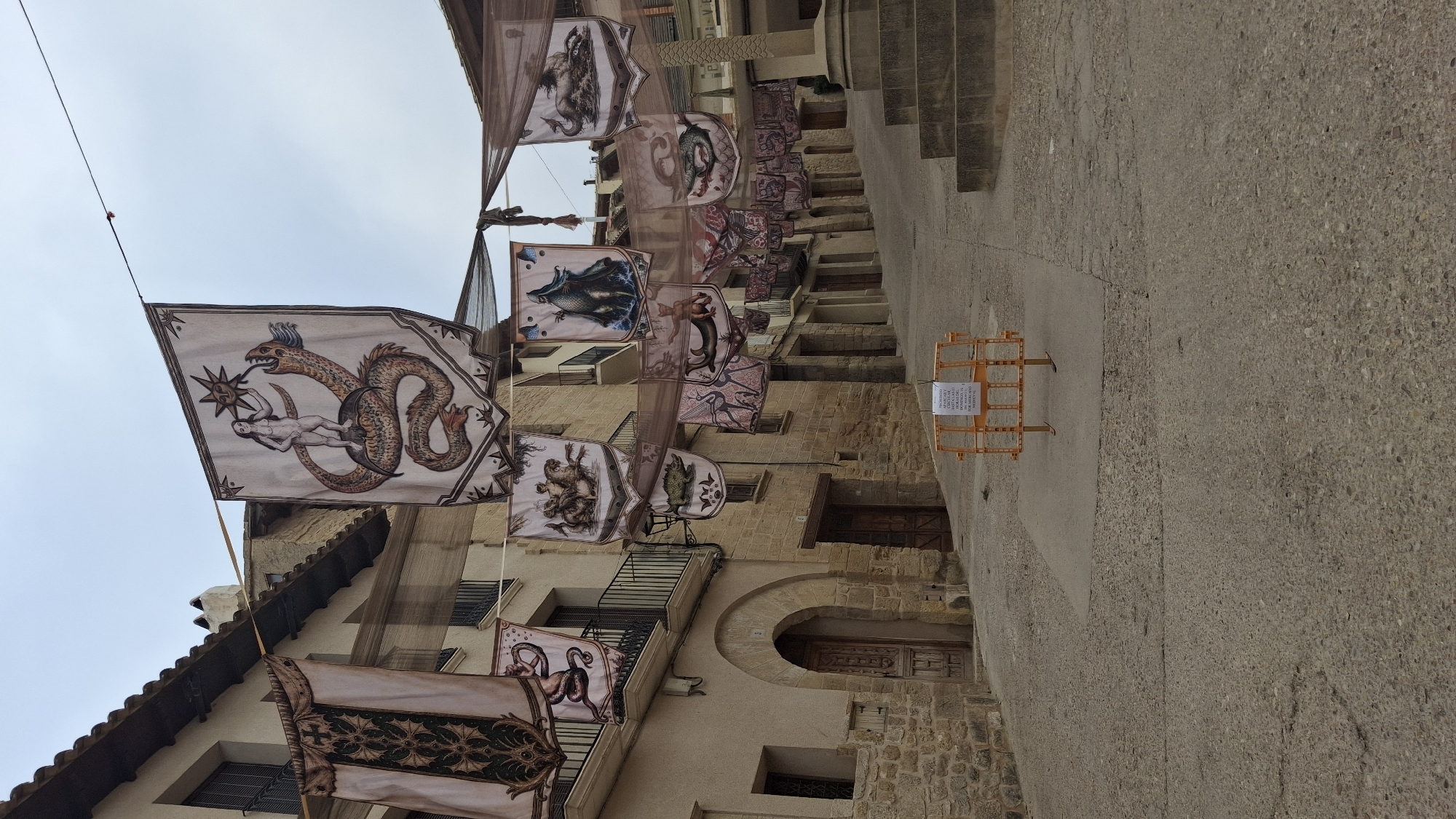 This image depicts a street scene in La Fresneda, Spain, adorned with numerous banners hung across the street. The banners, featuring medieval-style illustrations of mythical creatures and figures, are suspended from wires and a dark, sheer fabric netting. The street itself is paved with cobblestones and ascends a gentle slope, lined with old stone buildings with tiled roofs and balconies. Some banners display detailed imagery of dragons, knights, and other fantastical beings. In the foreground, a temporary orange construction barrier with a sign is placed on the side of the street. The sky is overcast, suggesting a cloudy day. The architecture and decorations hint at a historical or festive event taking place in the town.