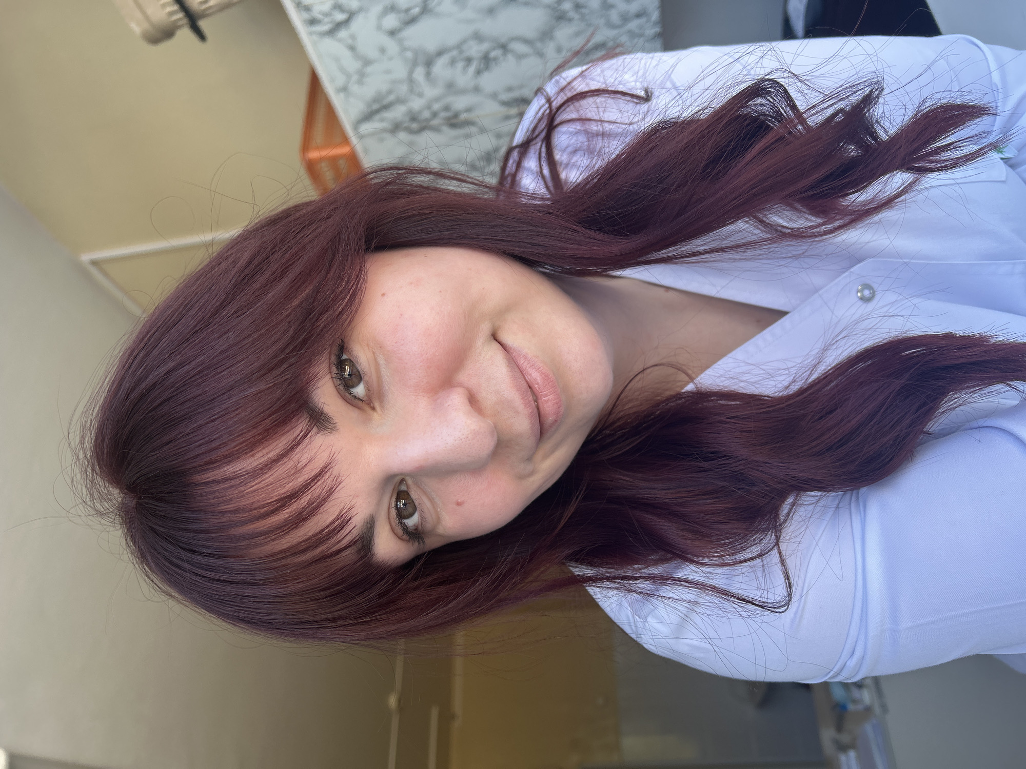 A woman with red-brown hair and bangs smiles at the camera. She is wearing a white shirt. The background includes a white countertop with a marble-like pattern. The location is possibly Фоки, Russia.