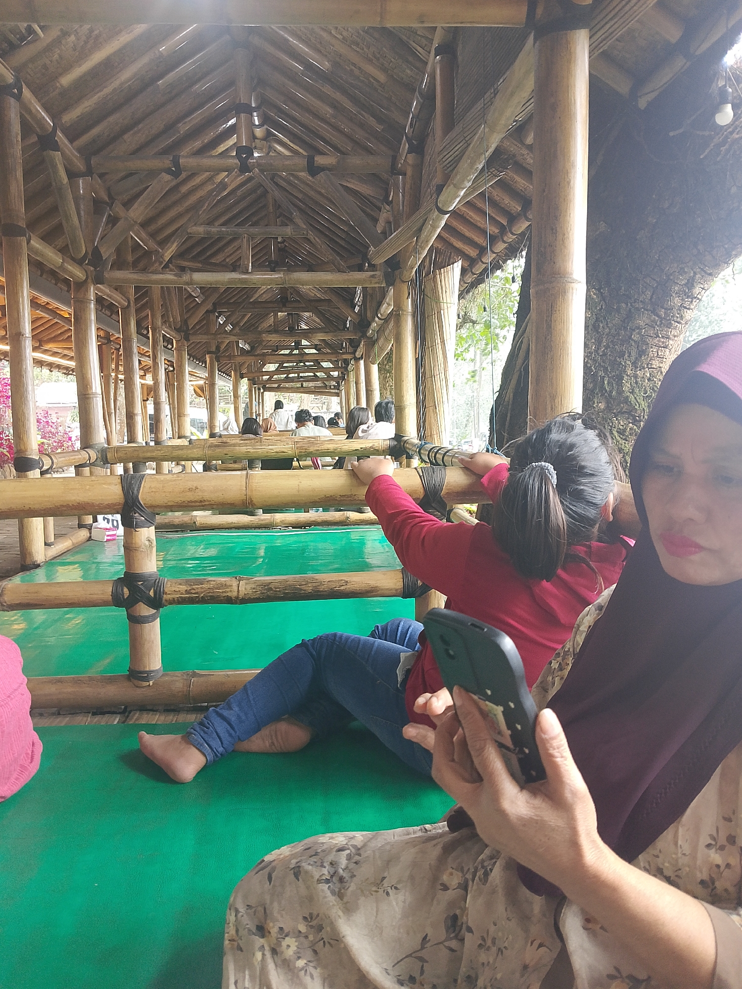 In Patengan, Indonesia, a person wearing a maroon hijab sits in the foreground, holding a black smartphone and looking towards it. Their hand, adorned with subtle veins, is visible in the lower right portion of the frame. To their left, a young girl in a red top and blue jeans is seated on a green mat, leaning against a bamboo railing. Her back is to the camera, and her hair is pulled back in a ponytail. She is reaching towards the railing. The scene unfolds within a covered structure with a bamboo roof and supports. In the background, several other individuals are seated, and the environment suggests an outdoor setting with greenery and a large tree trunk visible on the right. The overall impression is one of a relaxed social gathering within a rustic, open-air venue.
