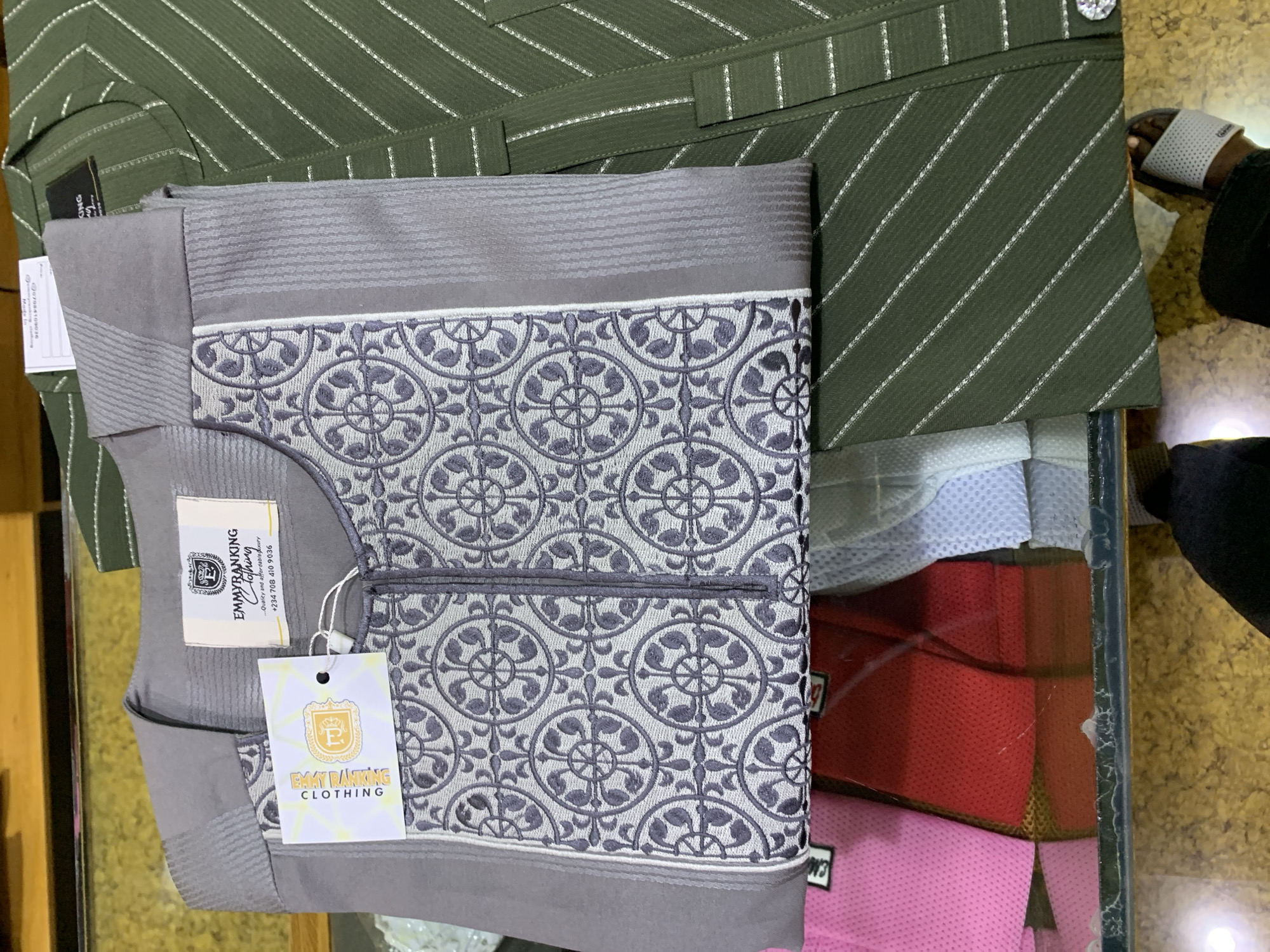 In a retail environment in Gura Top, Nigeria, a close-up shot displays two men's garments laid out on a glass surface. The foreground features a folded grey kaftan with intricate grey embroidery on the chest and a patterned yoke. A white tag with "EMMYRANKING CLOTHING" is attached to the kaftan. To the right, a green striped kaftan is partially visible, its vertical stripes subtly accented with thin, shimmering lines. The background reveals a shelf with other clothing items, including pink and red shirts, suggesting a retail display or inventory. The overall scene is brightly lit, focusing on the texture and detail of the garments.