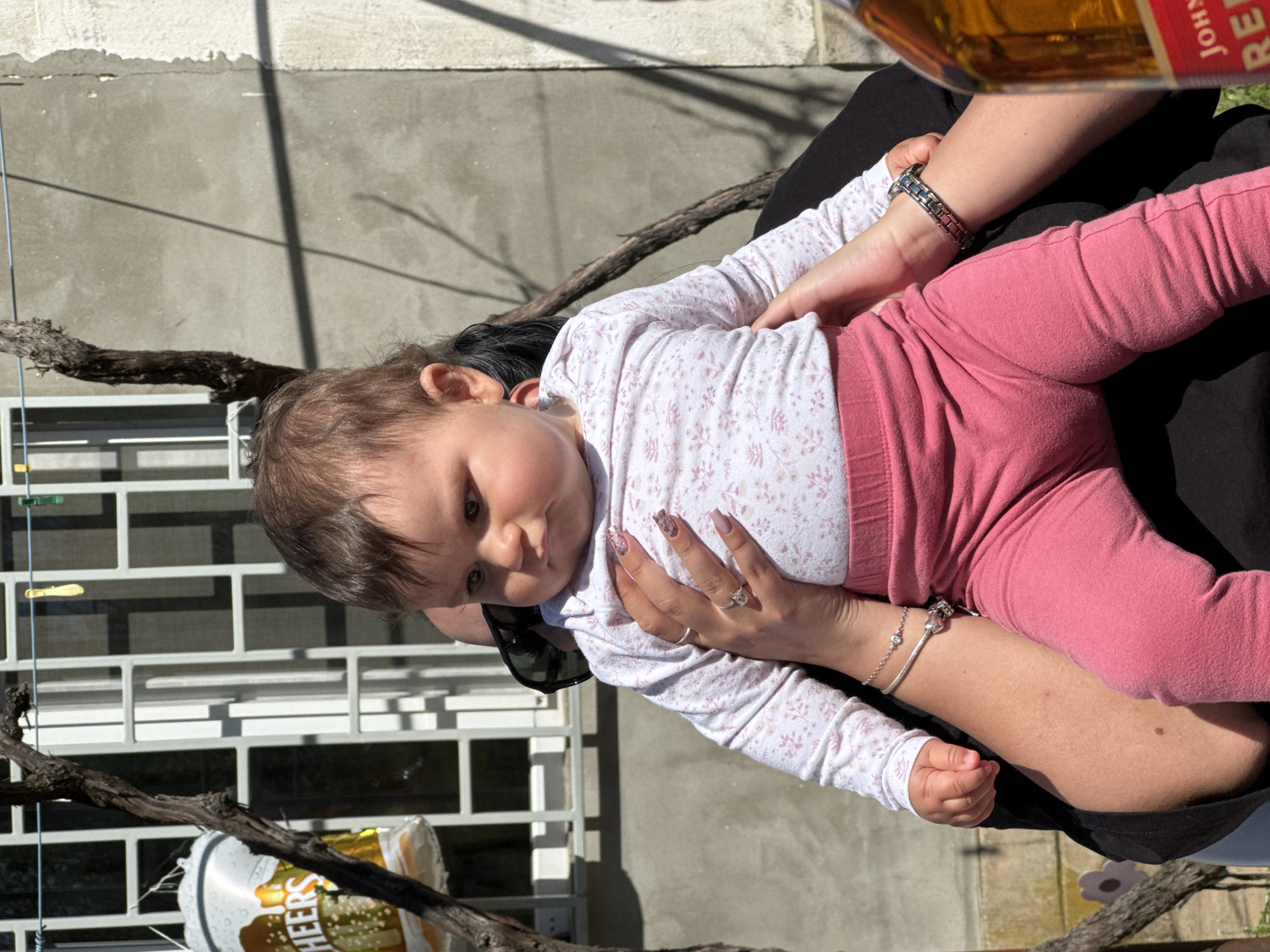 A baby wearing a white and pink floral shirt and pink pants is being held by an adult. The adult is wearing a silver watch and bracelet. The setting appears to be outdoors in Lom, Bulgaria. There is a white metal fence and a concrete wall in the background. A vine is visible near the fence, and a "CHEERS" balloon is partially visible in the lower-left corner. A bottle labeled "Johnnie Walker Red" is slightly visible in the upper right of the frame.