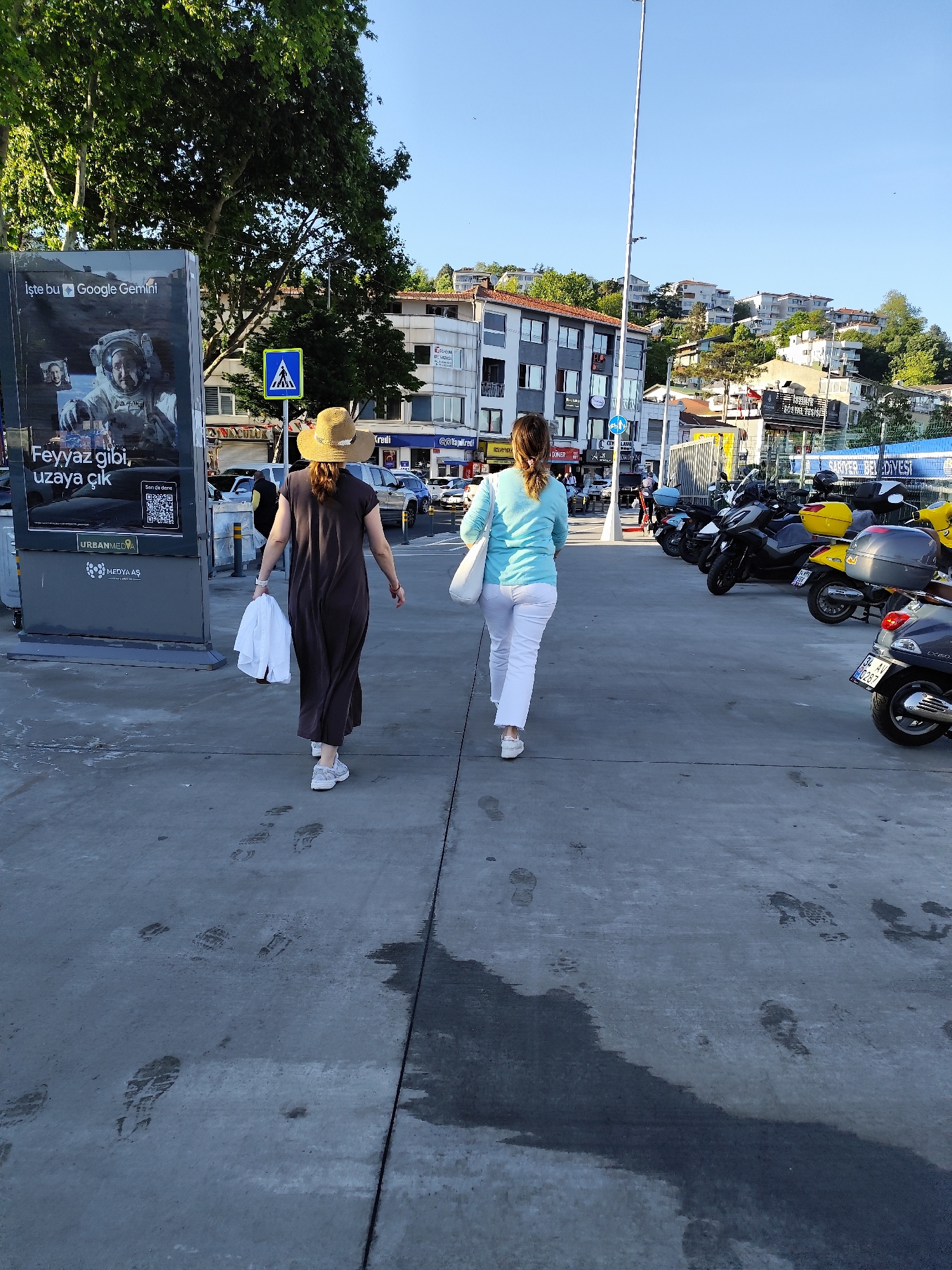 The image captures a scene in Sarıyer, Turkey. Two women are walking away from the camera on a paved area with footprints. The woman on the left wears a brown dress and a straw hat, carrying a white shirt. The woman on the right is dressed in a light blue shirt and white pants. To the right, there are several parked motorbikes and scooters.
In the background, there are several buildings and a street with light traffic. A sign for 'Sarıyer Belediyesi' is visible near the motorbikes. On the left, a billboard advertises Google Gemini. The weather appears to be sunny with clear blue skies.