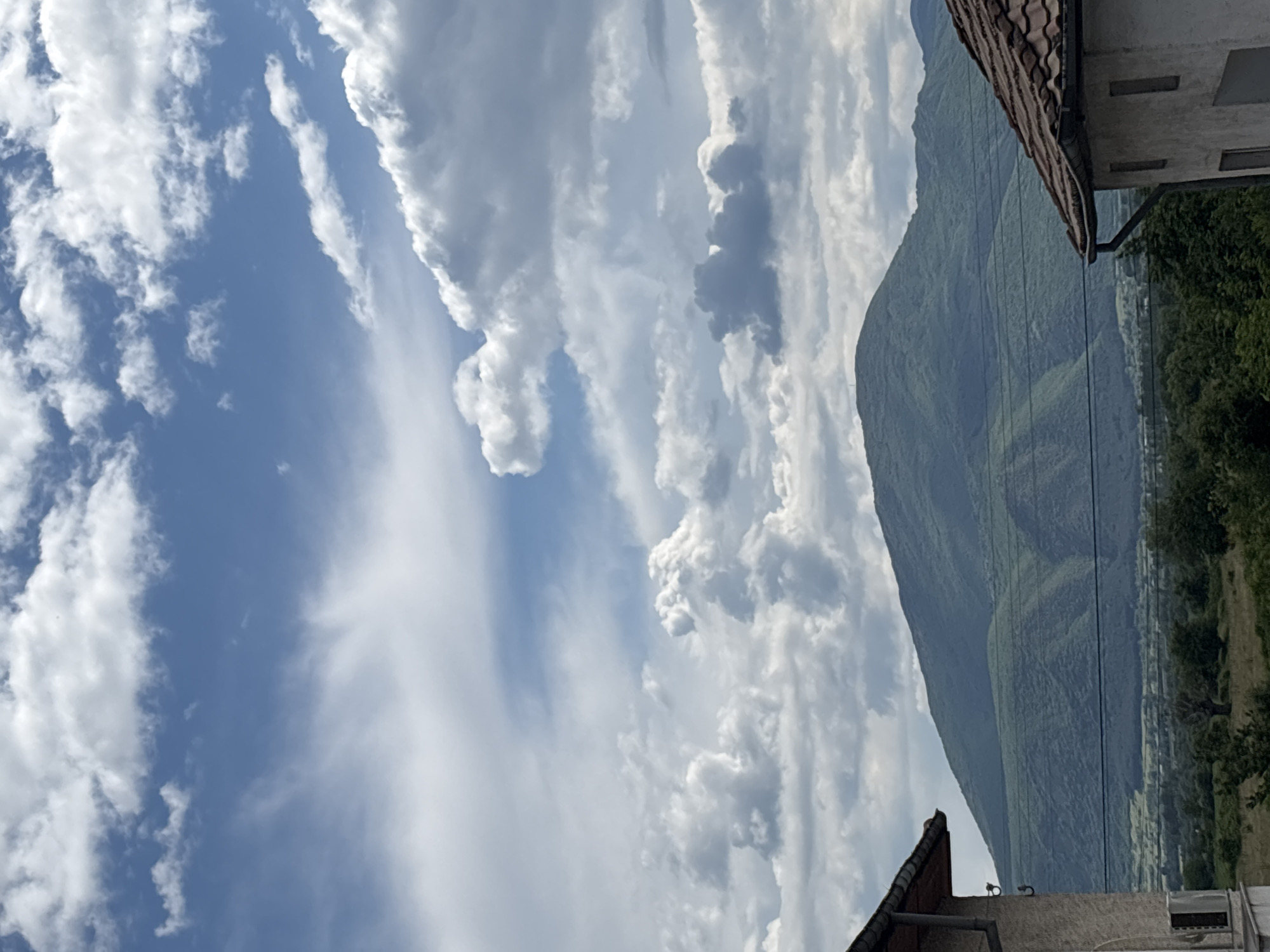 The image depicts a view in Garmen, Bulgaria, featuring a mountain range on the right side of the frame. The sky occupies a significant portion of the image, showcasing a mix of blue sky and white cumulus clouds. Building rooftops are visible at the top right and bottom of the frame, suggesting a residential area. Trees and possibly other greenery can be seen on the mountain's slopes and in the lower right, indicating vegetation. Power lines extend in front of the mountain.