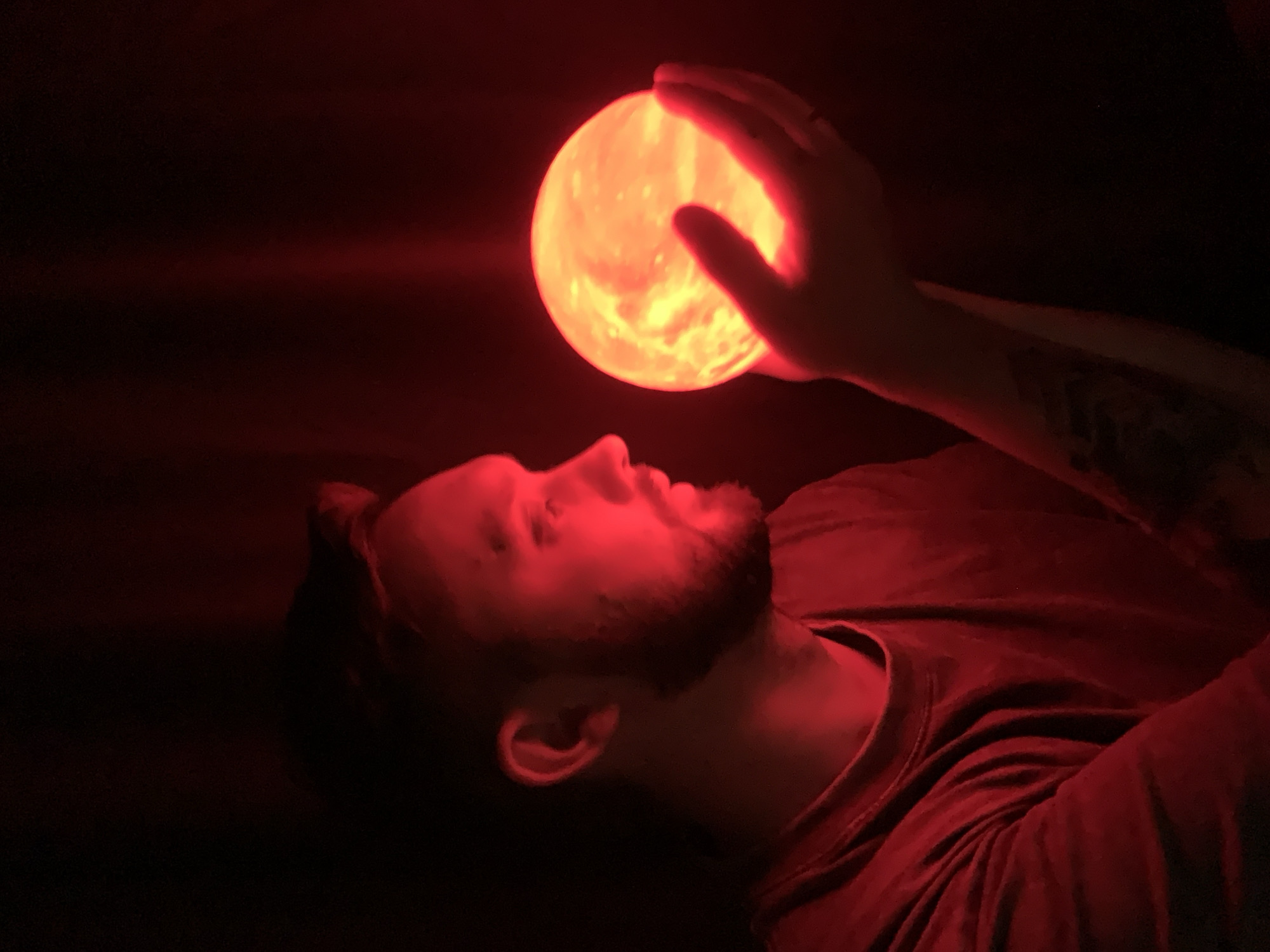A man lies on his back in near darkness, illuminated by a spherical, glowing red object held in his right hand above his face. The environment is dark; the location is indeterminable. The man has short brown hair, a beard, and is wearing a red t-shirt. The man has tattoos on his left forearm.
