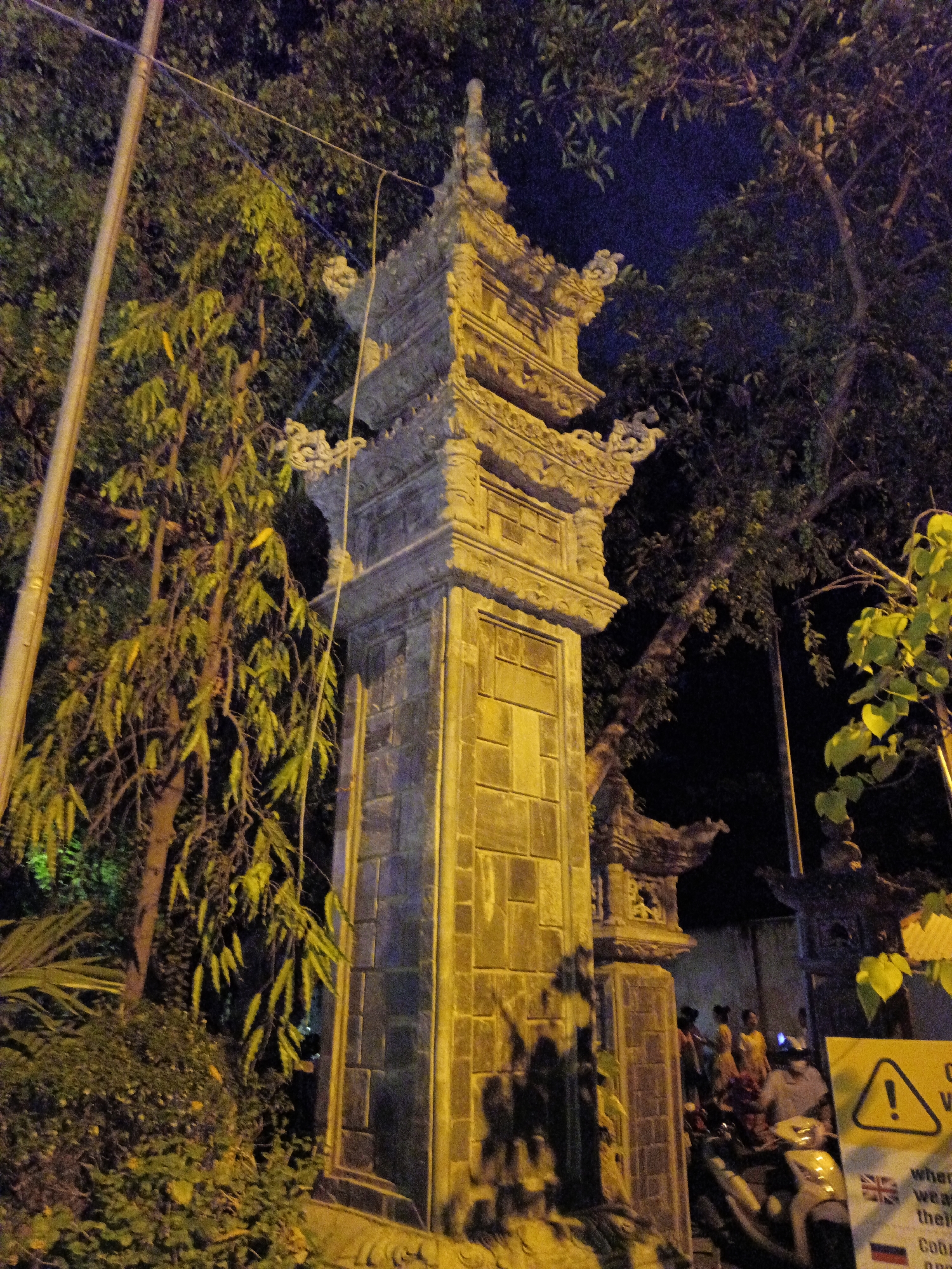The image shows a tall, ornate stone structure, possibly a temple gate or monument, illuminated by artificial light at night. It is located in Tay Nha Trang Ward, Vietnam. The structure is multi-tiered with decorative carvings on the edges. The base is made of stone blocks. Trees and dense vegetation surround the structure. Behind the structure, between two gates, there are several people. A sign with a warning symbol and text is visible in the foreground, next to a parked scooter. A pole is visible in the background, possibly a utility pole.