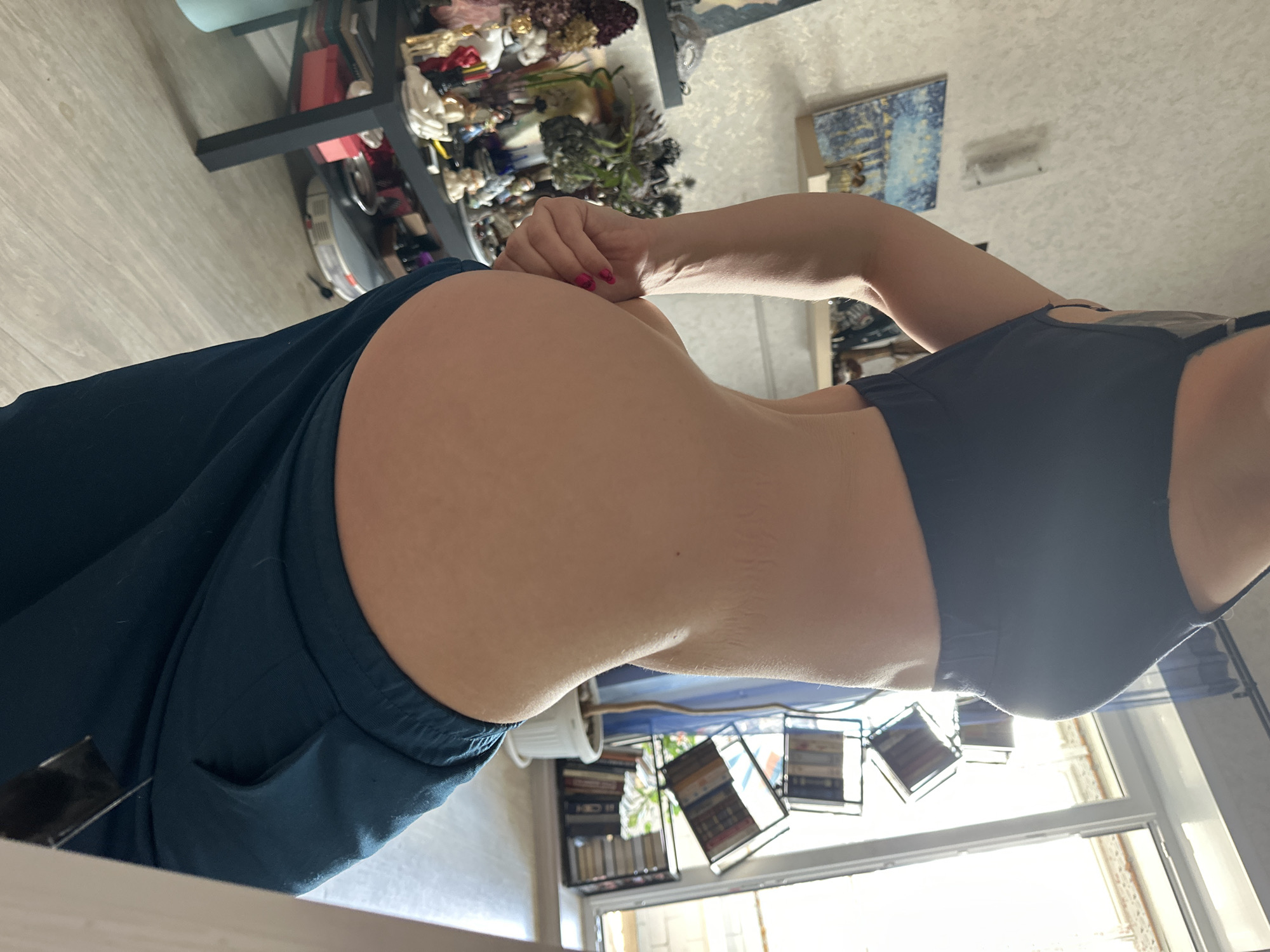A waist-level, indoor, side-view shot shows a person wearing a black sports bra and dark blue pants. The person is standing in front of a window with sunlight streaming through. In the background, to the left, there is a shelf with various objects on it, and a painting on the wall. Near the window, there are books and decorative frames. The person's right hand rests on their lower back. The location could be Vladimir, Russia, inferred solely from the user's profile.