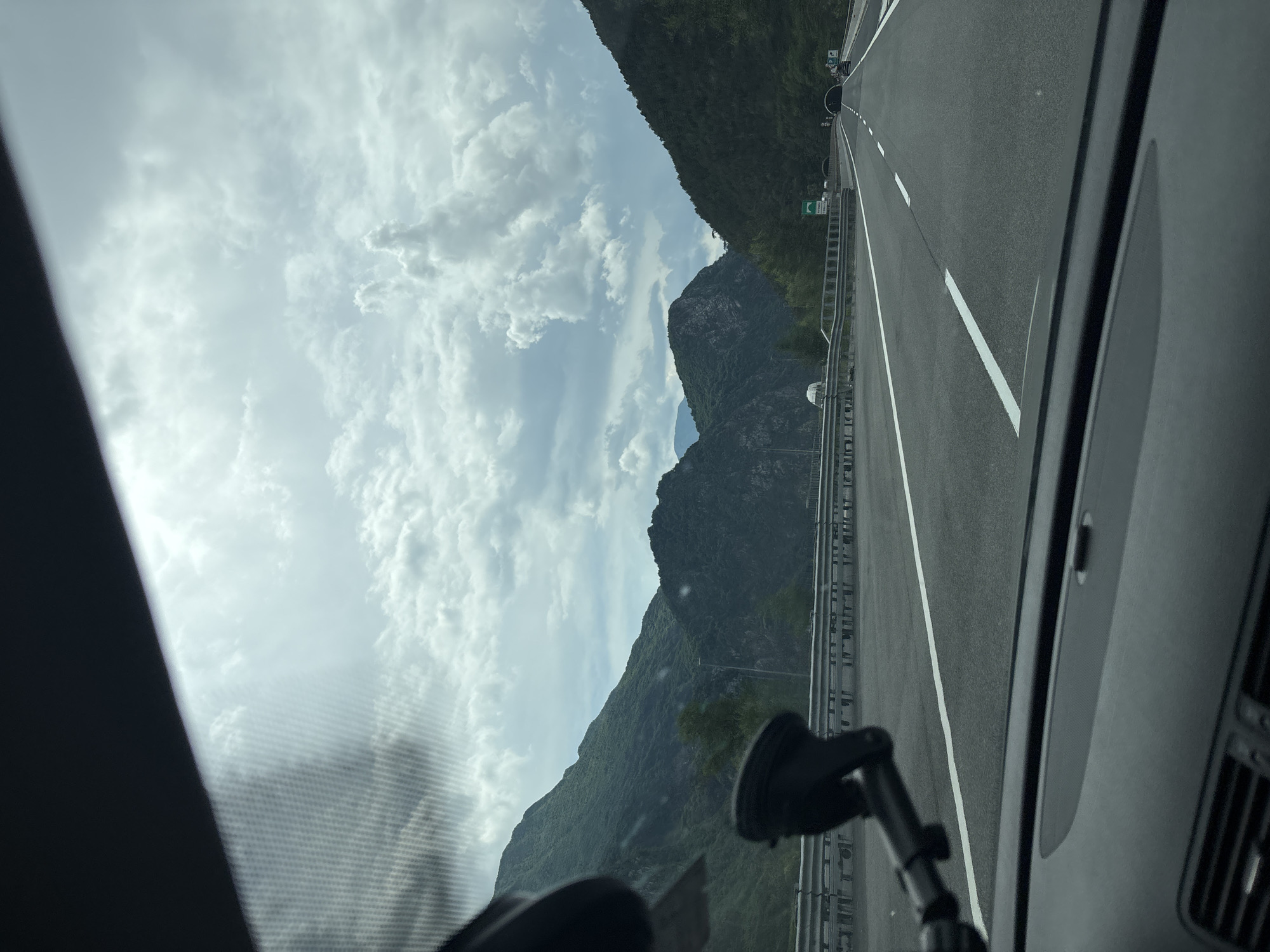 The image, taken from inside a vehicle, shows a highway in Moggio Udinese, Italy. The road has white lane markings and metal guardrails. Mountains are visible on the side of the road. The sky is cloudy. A mobile phone holder is attached to the windshield of the vehicle. The road leads to a tunnel.
