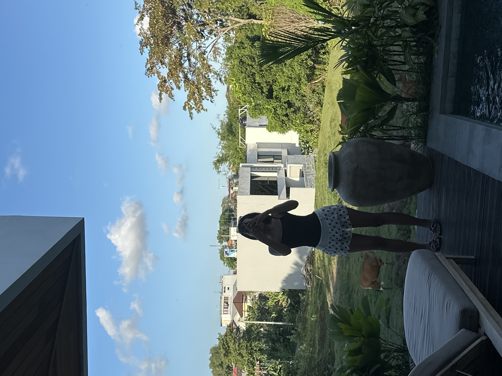 The image depicts an outdoor scene in North Kuta, Indonesia, featuring a woman standing on a wooden deck, taking a selfie with her phone. She is wearing a black tank top and a white skirt with black polka dots. To her left is a gray lounge chair, and to her right, a large, dark ceramic vase stands beside a swimming pool. In the background, a brown cow grazes in a grassy field adjacent to some modern buildings and lush green trees under a bright blue sky with scattered clouds. The overall environment suggests a residential or resort setting.