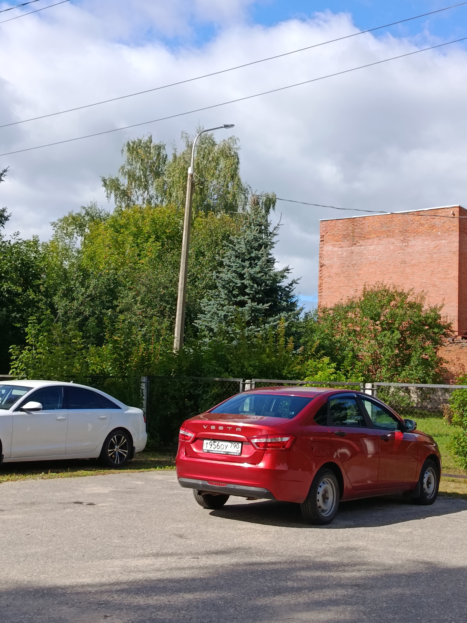 An outdoor shot captures a parking lot with two cars and a brick building under a partly cloudy sky. On the left, a white sedan is parked, partially obscured by a tree. To its right, a red sedan is parked, facing away from the viewer, with its license plate reading "T9560Y 790". The red sedan's taillights are visible, and a chain-link fence runs behind it, separating the parking lot from dense foliage and a tall brick building. A streetlamp stands near the center of the image, and power lines are visible against the sky. The scene is set in the city of Vereya, Russia.