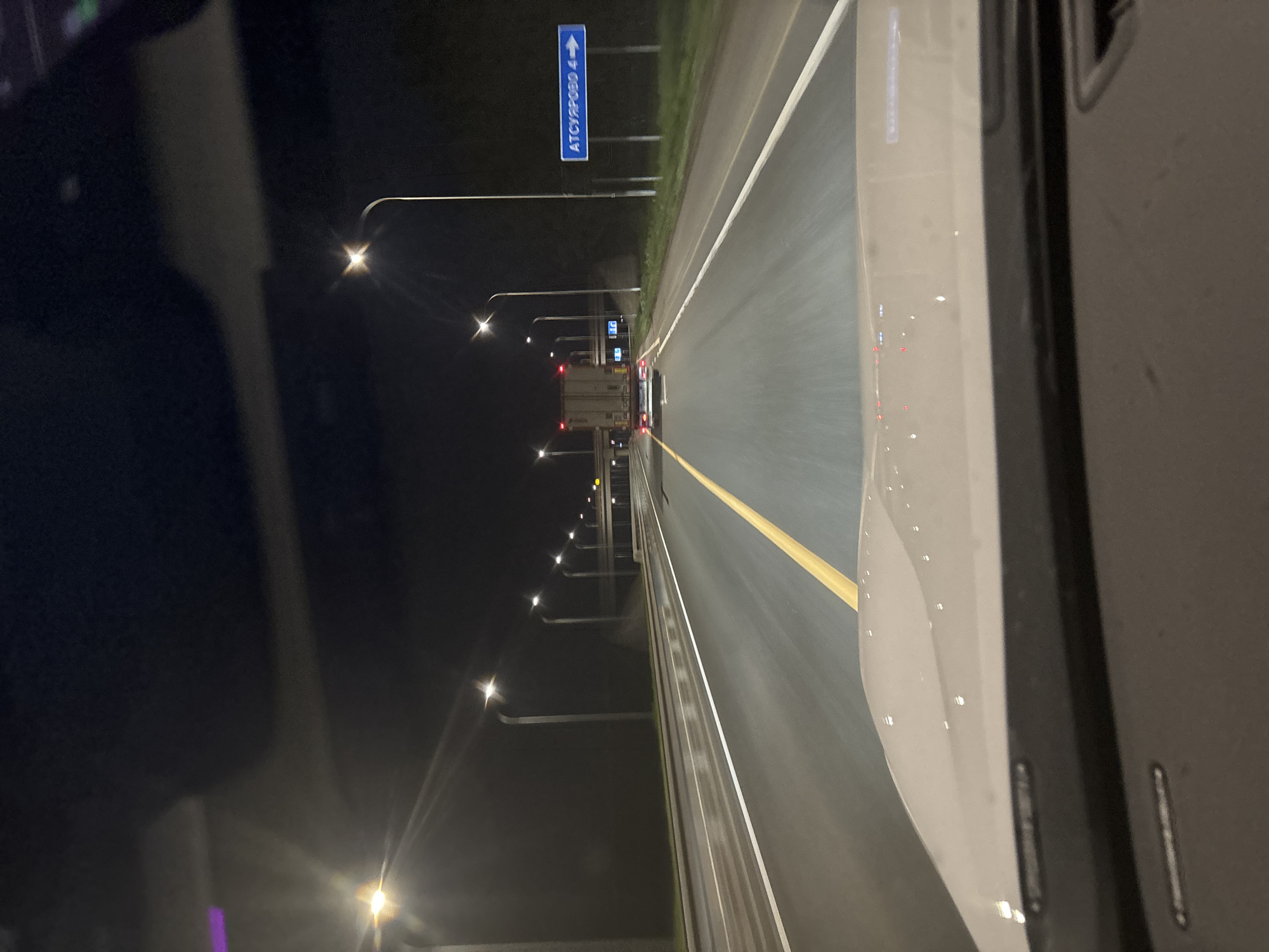 The image depicts a nighttime scene on a highway, likely in or near Таймурзино, Russia, based on the road sign indicating "ATCYPOBO 4" (likely referring to the direction of Ацырово, 4 kilometers away). The view is from inside a vehicle, looking forward. A truck is visible in the distance. Streetlights illuminate the road. Road markings include white lane dividers and a yellow line. The foreground shows part of the interior of the vehicle, specifically the dashboard and the car's body.