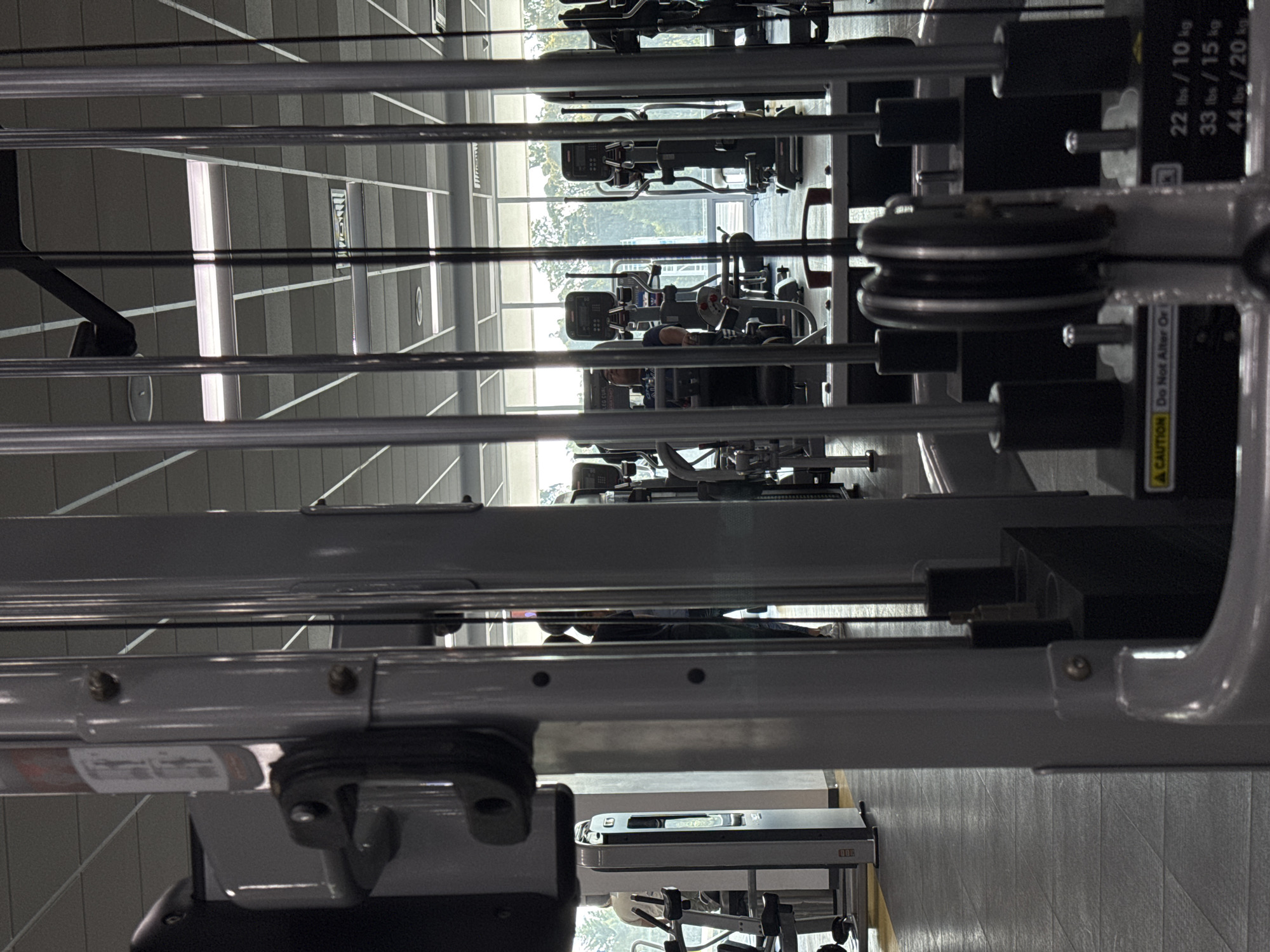 The image depicts a row of weight training machines in a gym setting. The machines are various models, and several people are using them. The gym has large windows, letting in natural light. The location is suggested to be in Heroica Puebla de Zaragoza, Mexico.