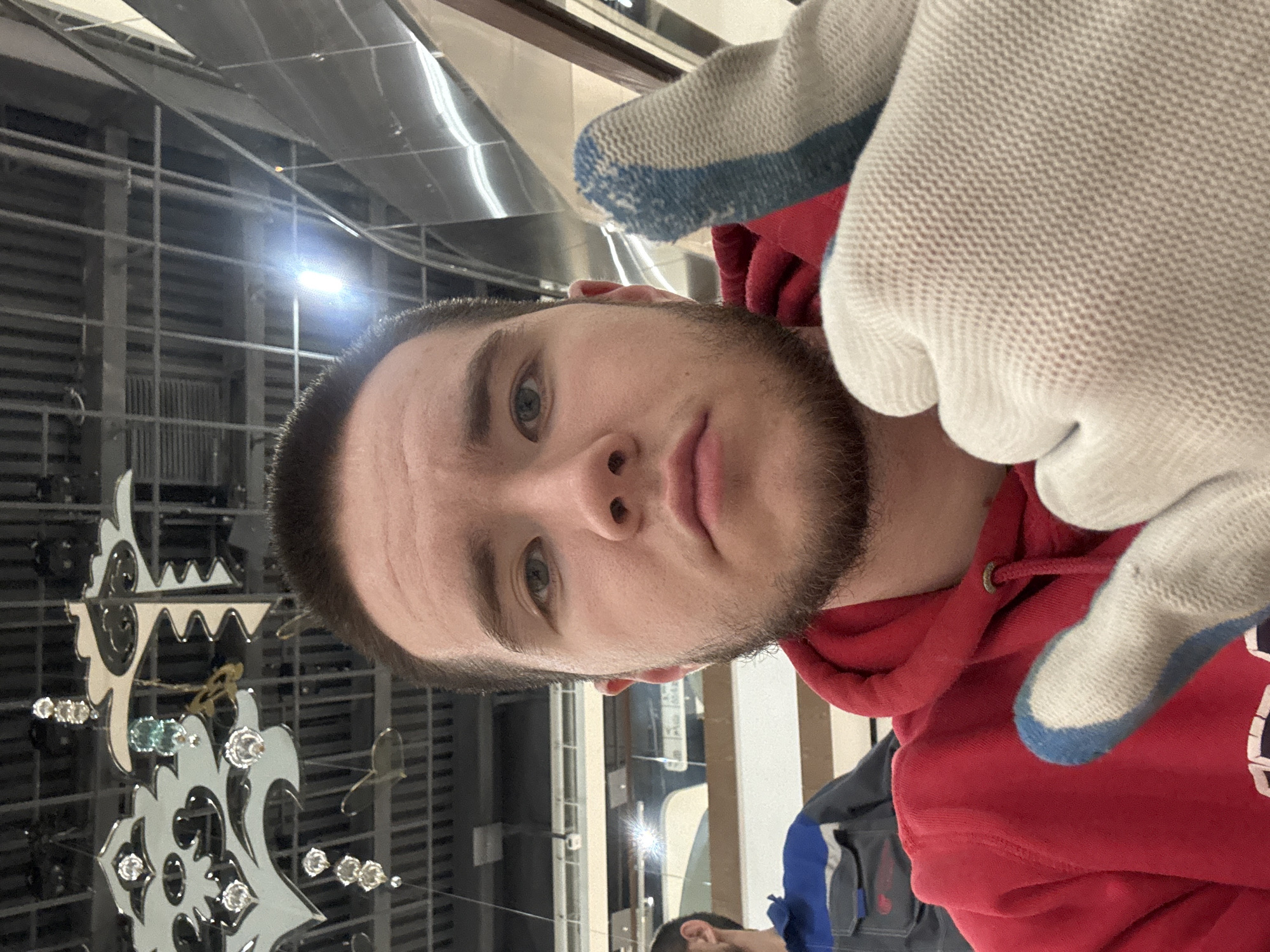 A rotated close-up portrait features a young man with light skin, short dark hair, and a neatly trimmed short beard, looking directly at the viewer with blue-grey eyes and a neutral expression. He is wearing a red hooded sweatshirt and white work gloves with prominent blue stripes and accents on the fingertips and wrist. His right hand, covered by a glove, is visible in the foreground, suggesting he might be holding the camera.
The background reveals an indoor setting with a combination of industrial and decorative elements. Exposed ceiling structures, possibly ventilation or metal framework, are visible, some with highly reflective surfaces. A bright artificial light source creates a glare in the mid-ground. To the man's left, an elaborate wall decoration or fixture, featuring layered, ornamental shapes with embedded clear stones or gems, adds an elegant contrast to the otherwise functional environment. A glimpse of another individual wearing a dark blue and grey jacket is visible in the lower background. The scene appears to be within a commercial or public building, possibly a showroom, retail space, or an area undergoing installation or maintenance, located in Kazan, Russia. No visible text is present.