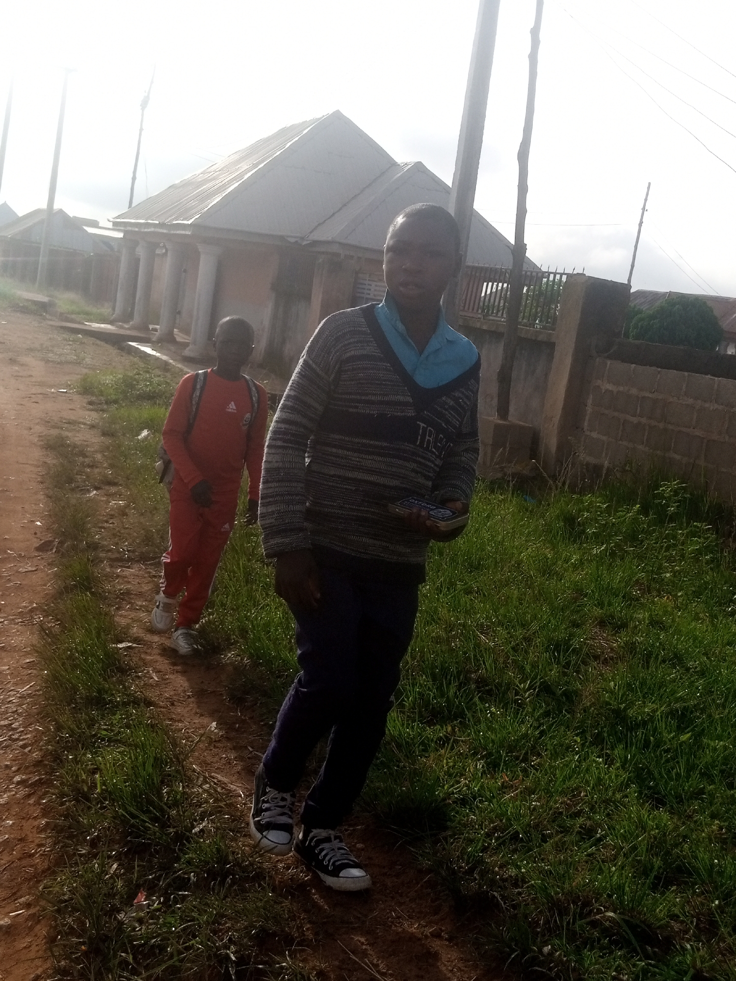 Two young males are depicted in an outdoor environment, possibly on a dirt road. The individual in the foreground is wearing a striped sweater over a light blue collared shirt and dark pants, with black and white sneakers. He is holding a rectangular object, possibly a book or electronic device, in his right hand. The second individual, positioned behind and to the left of the first, is wearing a full orange tracksuit and white sneakers, with a backpack on his shoulders. He is looking towards the ground. The background shows buildings with corrugated metal roofs and pillars, along with a brick wall and lush green vegetation. A utility pole with wires is also visible. The lighting suggests it is daytime, with the sun positioned high in the sky, causing some glare. The overall scene suggests a common street or residential area in a Nigerian city like Russo.