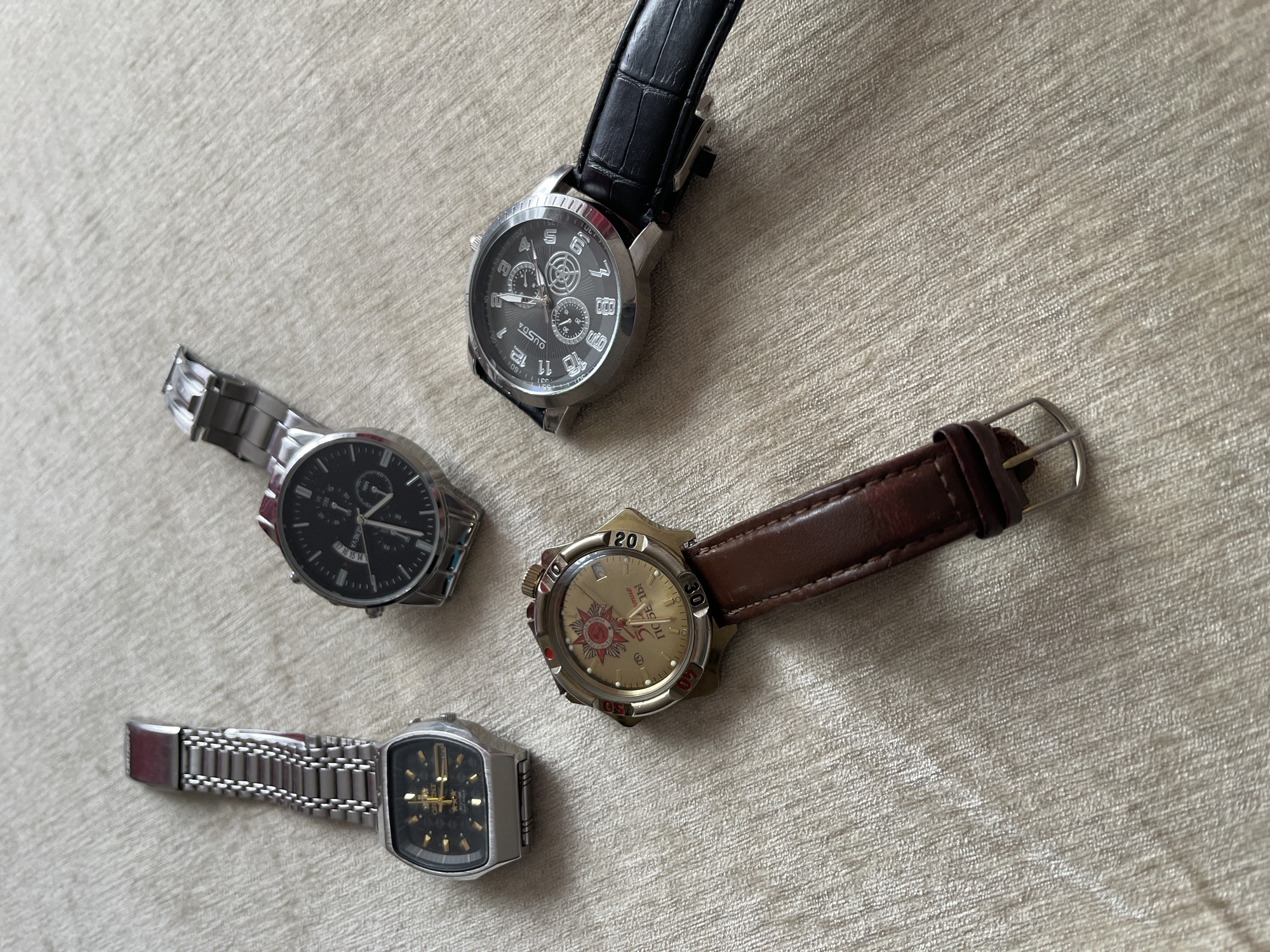 The image shows four wristwatches lying on a textured surface. The watches are positioned at different angles.
From top to bottom, the first watch has a silver case and a black leather band. Its dial features multiple subdials and prominent numeral hour markers. The name "OUSDA" is visible on the dial.
The second watch has a silver metal case and a silver metal band. Its dial is black with silver-colored hour markers and hands, and a date window.
The third watch has a gold-colored case and a brown leather band. Its dial has the word "ПОБЕДЫ" and a star.
The fourth watch has a rectangular silver case and a silver metal band. Its dial is black with gold-colored hands and hour markers.