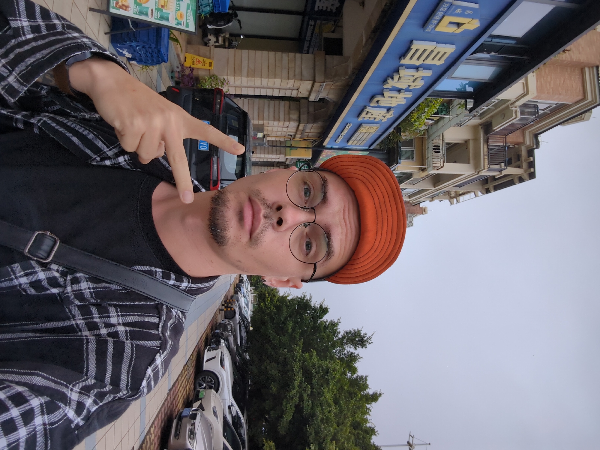 A man wearing an orange hat and round glasses poses for a selfie in Shunde District, China. He makes a peace sign with his fingers. The man wears a black and white plaid shirt over a black t-shirt. A black bag strap crosses his torso. In the background, there are cars parked along the side of the street and trees. Buildings and signage are also visible, including a sign with Chinese characters. A black car with the license plate "VO" is behind the man's hand.