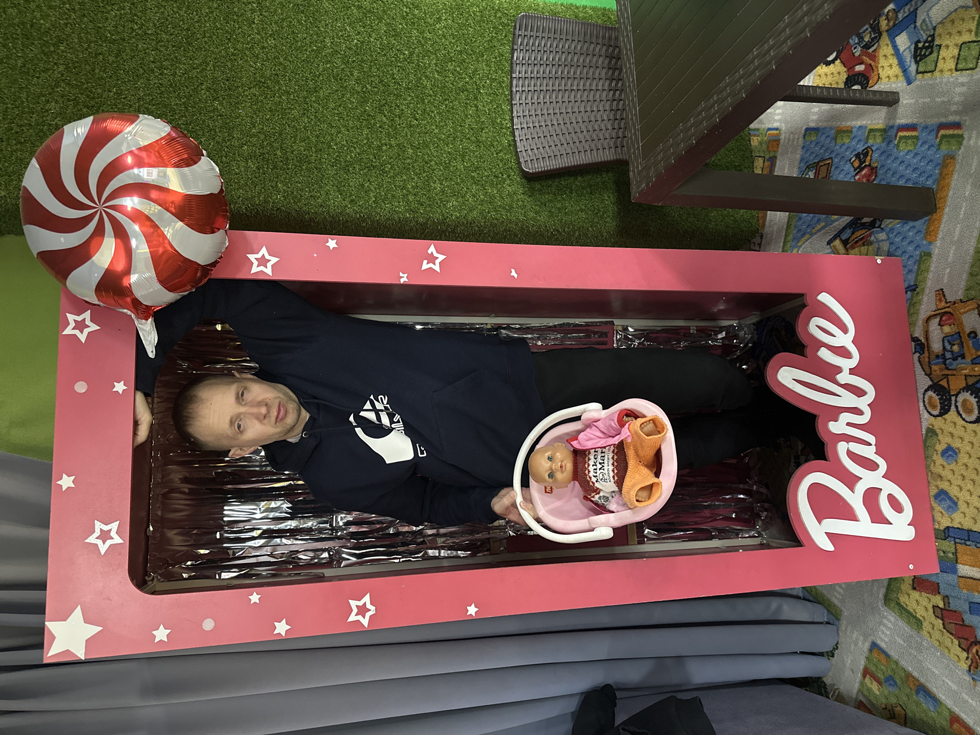 A man is lying inside a pink "Barbie" box on what appears to be a cushioned floor in what might be a playroom in Glazov, Russia. The box is decorated with stars and the "Barbie" logo. The man is holding a pink bucket containing a doll and some clothing. Next to the box is a red and white swirled balloon. There is furniture visible in the background, including a brown wicker chair and a table. Part of the floor is covered with what appears to be green artificial turf, and another part has a rug with cartoon-style vehicle graphics.