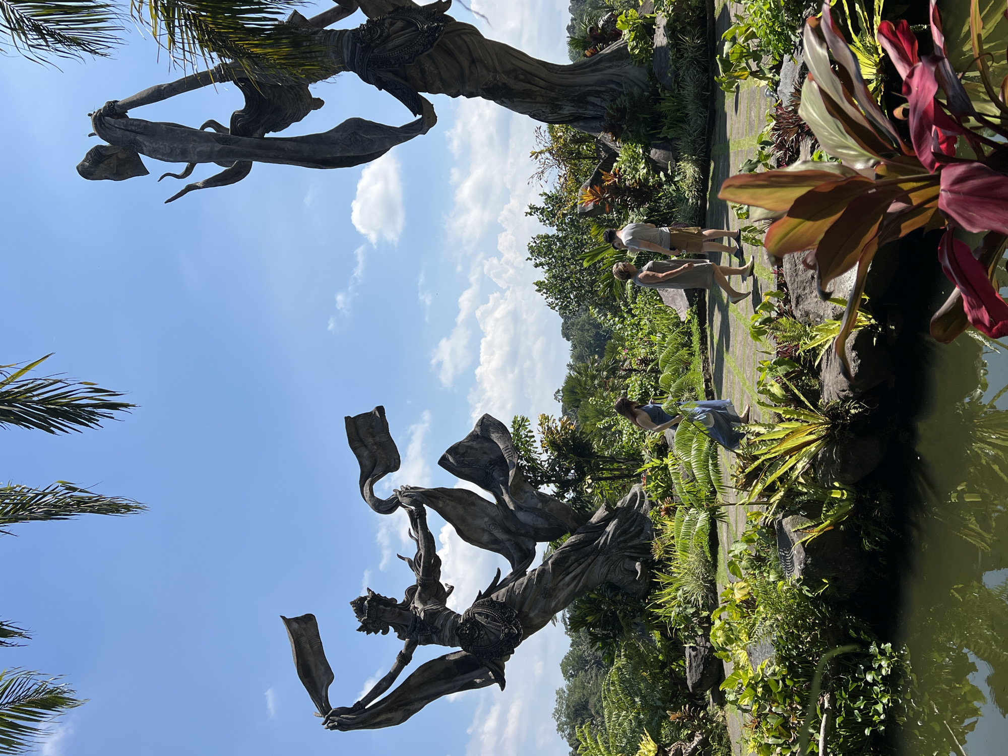 The image depicts a garden setting in Kedewatan, Indonesia, featuring a large stone sculpture of a woman with flowing fabric. The sculpture is positioned against a backdrop of a blue sky with scattered clouds. Below the sculpture, the scene transitions into lush vegetation, including various plants and trees. Two pairs of people are observing the scene: one pair is standing and looking in the distance and the other is standing by the pond. A pond reflects the sculpture and greenery, creating a symmetrical effect. The overall composition suggests a cultural or artistic attraction within a natural environment.