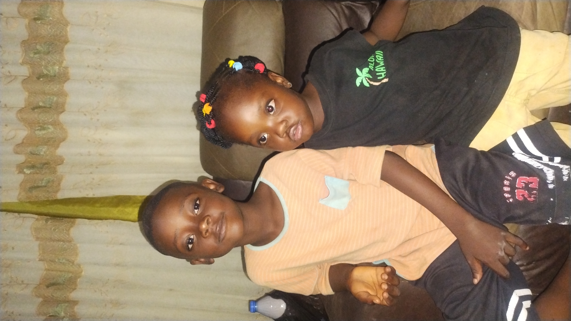 Two dark-skinned children are seated side-by-side on a brown leather-like sofa in an indoor setting in the city of Jos, Nigeria. The background consists of light-colored curtains with a textured pattern. A vertical strip of green fabric is visible on the far left.
The child on the left, a boy with short dark hair, wears an orange horizontally striped long-sleeved t-shirt with a light blue chest pocket, and dark shorts with white stripes and a red "SPORTS 23" logo. His left hand is open, palm up, with fingers slightly curled. He looks directly at the camera with a neutral expression.
The child on the right, a girl with dark hair styled in braids adorned with red, yellow, and blue beads, wears a black t-shirt featuring a green palm tree and the text "ALOHA HAWAII." She wears light-colored shorts. She looks upwards and to the right of the camera with a slightly downturned mouth.
A plastic bottle with a blue cap and dark liquid is visible on the sofa to the left of the boy.