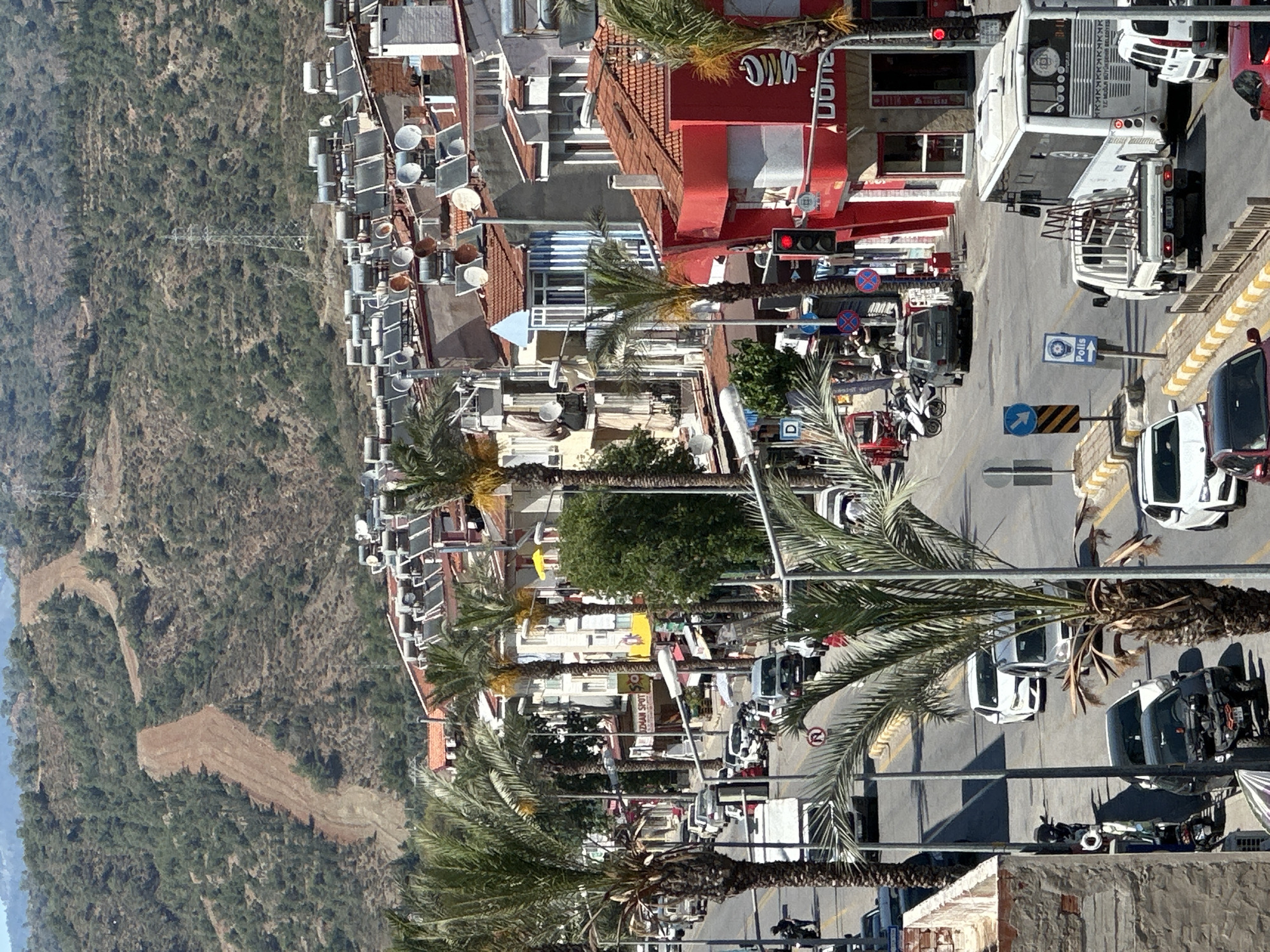The image presents a high-angle view of a bustling street in Fethiye, Turkey, bathed in bright daylight under clear skies. The street is flanked by multi-story buildings, many featuring red-tiled roofs, flat roofs laden with solar water heaters, and satellite dishes. Numerous tall palm trees line the street, casting shadows and adding to the Mediterranean atmosphere.
The road itself is active with traffic, including cars, motorcycles, a white bus, and a white pickup truck with a rack. Some vehicles are parked, while others are in motion, and a traffic light is clearly visible showing red. In the distance, a large, green, and rocky hillside rises, featuring a prominent communication tower and a winding dirt path.
Visible text includes "DÖNER" on a red building, likely indicating a restaurant. The side of the white bus displays "FETHİYE OTOGAR" (Fethiye Bus Terminal). A street sign points with the text "Polis" (Police), and another sign appears to read "ÖZDEM SPOT." Various standard traffic signs are also present. The scene captures the ordinary daily life and activity of a sunny day in the town.