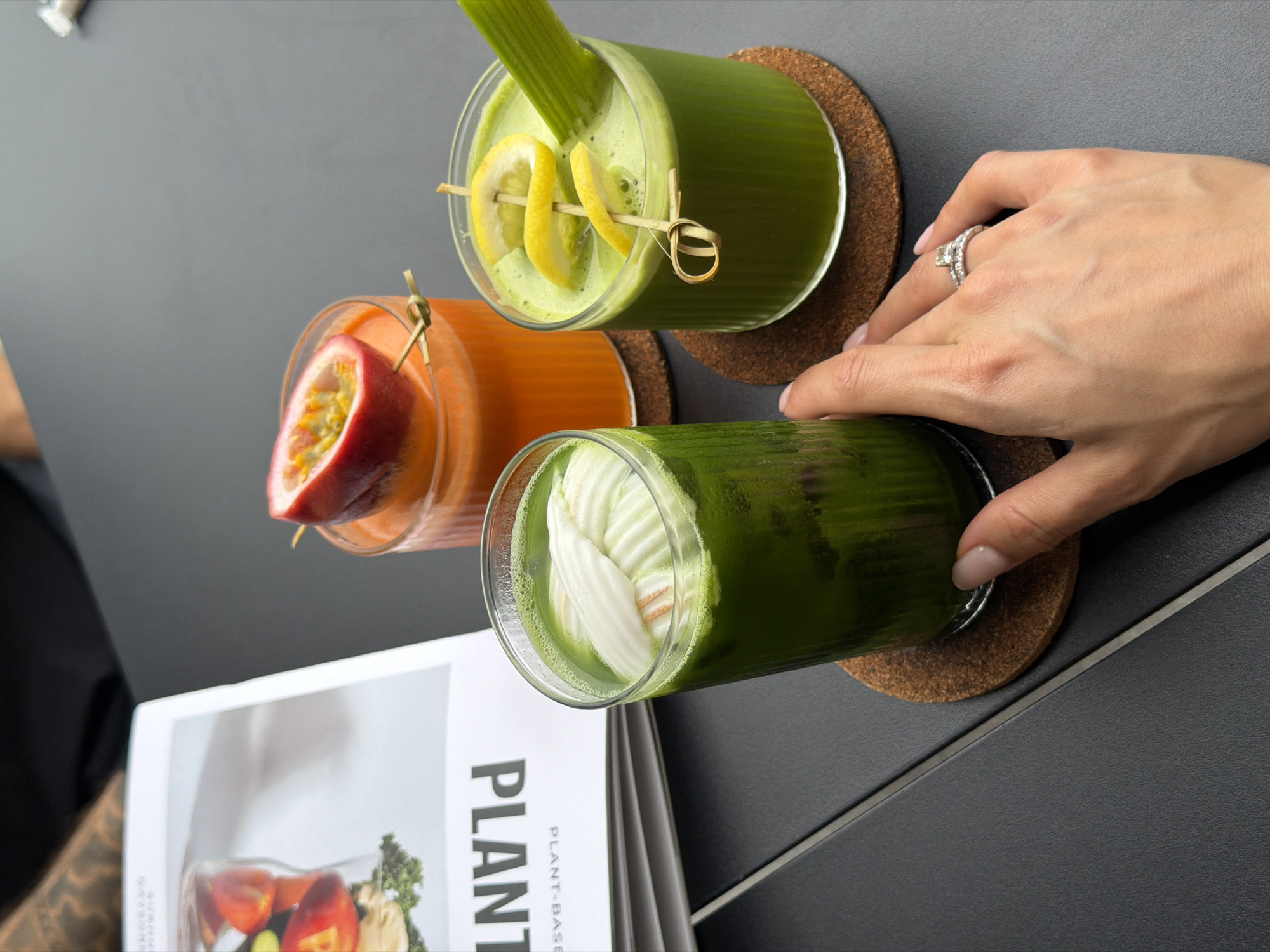 A high-angle shot presents three drinks on a dark gray surface, likely a table, indoors. Starting from the top, the first drink is green with a celery stalk and lemon garnish on a wooden skewer, sitting on a cork coaster. Below it, an orange drink is adorned with a passion fruit slice on a skewer. The third drink, lower in the frame, is green and decorated with a white vegetable garnish. It also sits on a cork coaster. A hand with a diamond ring rests on the edge of the frame near the top drink. In the lower left corner, the cover of a book or magazine with the word "PLANT" is visible. Given the context, the drinks are likely at a cafe or restaurant in Choeng Thale, Thailand, specializing in plant-based beverages.