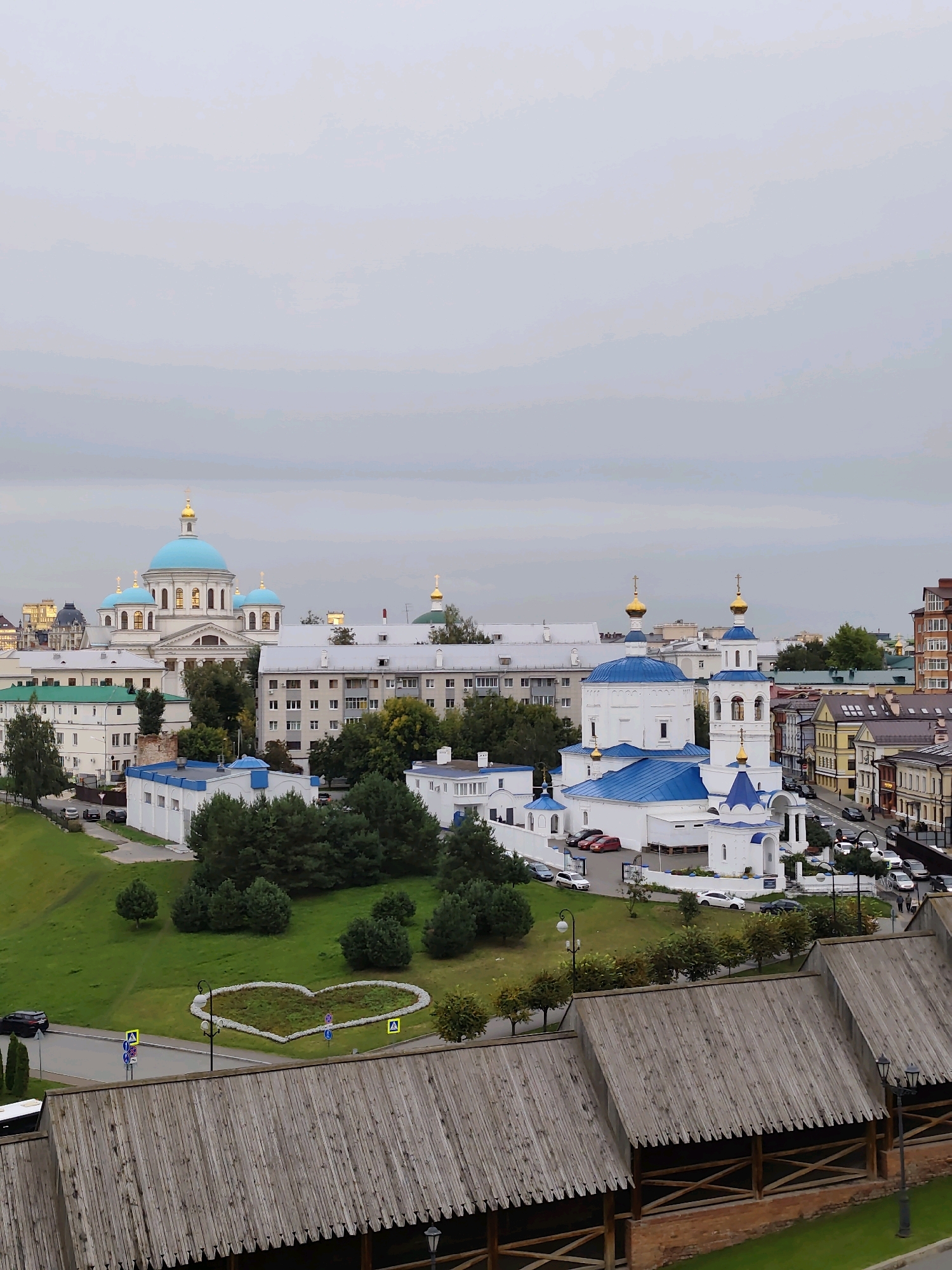 The image shows an aerial view of the Kremlin in Kazan, Russia, on a cloudy day. In the foreground, a wooden roof structure with visible beams and planks dominates the bottom portion of the frame. Behind this structure, a lush green lawn is visible, featuring a heart-shaped flower bed filled with colorful flowers. Several trees are scattered across the lawn, and a road with cars and pedestrian crossings runs alongside it.
Further back, several white buildings with blue and golden domes stand out. The most prominent is a large church with multiple domes, characteristic of Russian Orthodox architecture. Adjacent to it are other buildings with similar architectural features, including a bell tower. In the background, more modern buildings and residential structures are visible, interspersed with trees. The overall impression is a blend of historical religious sites and urban development. The sky is overcast, casting a soft light on the scene. There are no visible people in the image.