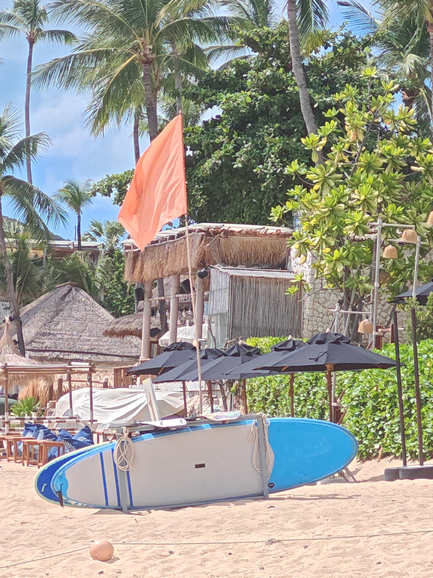 A bright orange flag is prominent in the upper center of the image, attached to a pole. In the foreground, two stand-up paddleboards are laid out on the sandy beach, one partially covering the other. The top board is white with blue stripes, and the bottom board is primarily blue. Ropes are used to secure the boards. Behind the paddleboards, several black umbrellas are open, providing shade. Further back, there are thatched-roof structures, palm trees, and dense green foliage. The sky is visible in the background with some clouds. The scene appears to be a beach resort area in Baan Bo Phut, Thailand.