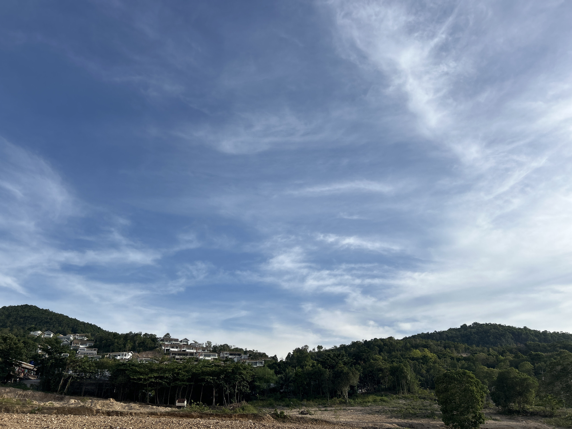 The image shows a daytime scene in Baan Wang Ta Kien, Thailand. A clear blue sky with thin, streaky cirrus clouds dominates the upper two-thirds of the image. In the lower third, a sloping hillside is covered in lush green trees. Dwellings are clustered near the top of the hill on the left. In the foreground, there is light brown, dirt land. The ground is mostly flat and seems dry. There are hints of disturbed earth. The photograph has been taken in daylight.