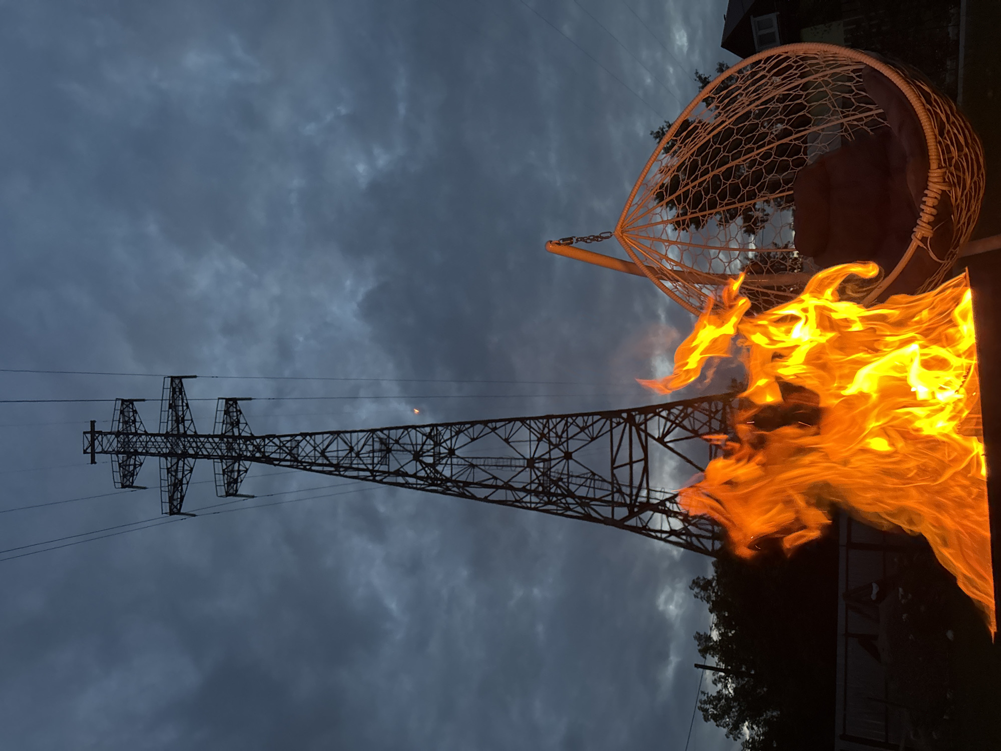 The image depicts a nighttime scene in Chaikovsky, Russia, featuring a bonfire in the foreground and a large metal power line tower in the background. The sky is overcast with dark clouds, illuminated by the glow of the fire and a faint light from a distant source, possibly the moon or a streetlamp. A white woven hanging chair with a dark cushion is positioned to the right of the fire, creating a cozy atmosphere. Silhouetted trees and a fence are visible behind the fire, suggesting a residential or backyard setting. The overall impression is one of a serene, outdoor evening in a semi-urban environment.