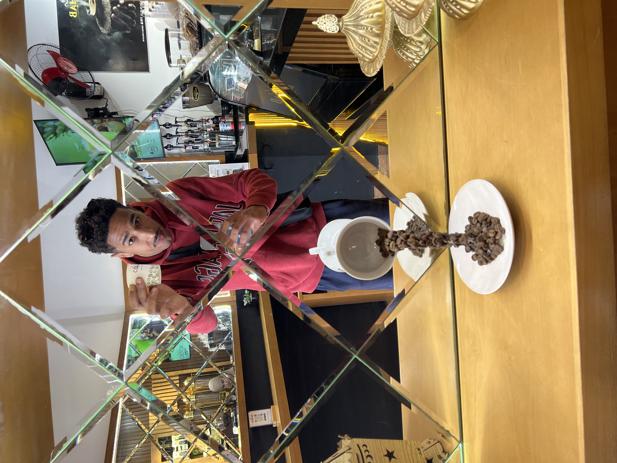 The image depicts the interior of a modern coffee shop in Tanger, Morocco, viewed through a decorative wall composed of diamond-shaped mirror tiles. In the foreground, on a light wooden counter, a white ceramic coffee cup appears suspended mid-air, with a stream of roasted coffee beans seemingly pouring from it onto a small white saucer below. To the left of this installation is a wooden decorative lantern-like object, and to the right, a golden ornate covered dish.
A male subject with dark, curly hair and a mustache, wearing sunglasses on his head and a red hooded sweatshirt, stands in the midground. He is holding a small white disposable cup with green leaf patterns and Arabic script in his right hand, extending it towards the camera. He wears a black shoulder bag and is looking directly at the viewer.
The background, partially obscured and reflected by the mirrored tiles, reveals the coffee shop's operational area. Behind a dark counter, a female barista in a light-colored hijab is visible. Various coffee machines, grinders, and syrup dispensers are arranged on the counter. Wall-mounted screens display promotional content, including a "DETOX 35%" sign and a QR code labeled "UNSUM." A black wall fan with red blades is mounted on the upper right wall, near posters featuring text like "ARABICA." The counter area is illuminated by yellow strip lighting.