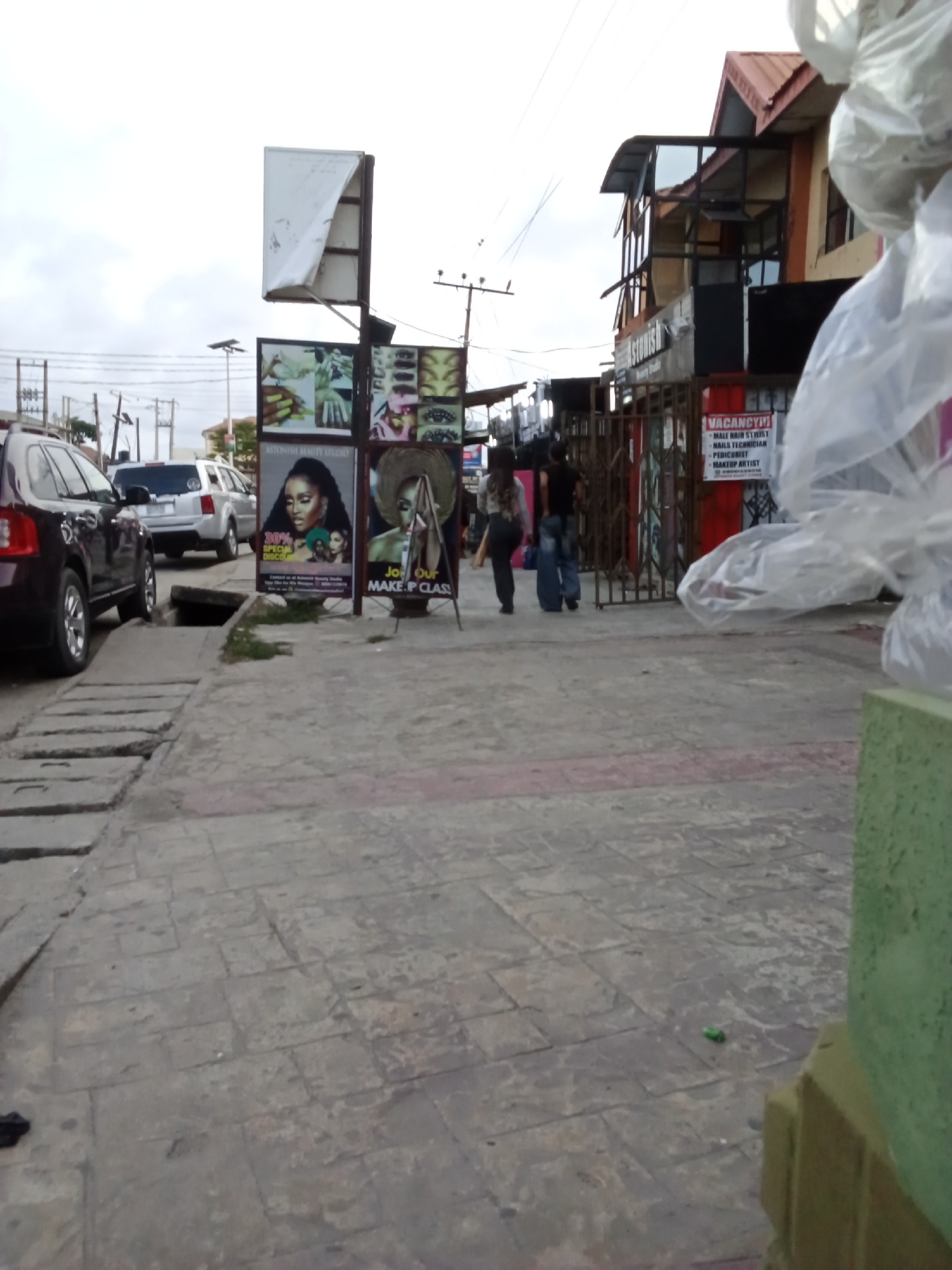 This outdoor street scene, likely captured in Ado, Nigeria, depicts a bustling urban environment on an overcast day. The foreground features a paved sidewalk made of concrete slabs, with a dark SUV parked parallel to the curb on the left. A concrete drain or gutter structure runs alongside the parked vehicle.
In the mid-ground, two people are seen walking away from the camera on the sidewalk, passing by several large advertising boards. One sign prominently displays "ASTONISH BEAUTY STUDIO" with an offer for "30% SPECIAL DISCOUNT." Another sign adjacent to it encourages viewers to "JOIN OUR MAKE-UP CLASS," featuring an image of a person wearing a straw hat. Above these, a tall, white sign appears partially damaged or dislodged.
To the right, a red and white "VACANCY!!!" sign is affixed to a metal gate or fence, listing openings for a "MALE HAIR STYLIST," "NAILS TECHNICIAN," "PEDICURIST," and "MAKEUP ARTIST." Behind this gate, a multi-story building with a red-tiled roof and visible window frames is partially obscured. Overhead utility poles and numerous power lines crisscross the bright, cloudy sky, further defining the urban setting. On the far right, clear plastic bags are visible, partially obscuring a green and beige structure. A silver SUV is parked further down the street behind the dark SUV. A small green object rests on the pavement in the lower right. The scene suggests a commercial area with ongoing pedestrian and vehicular activity.