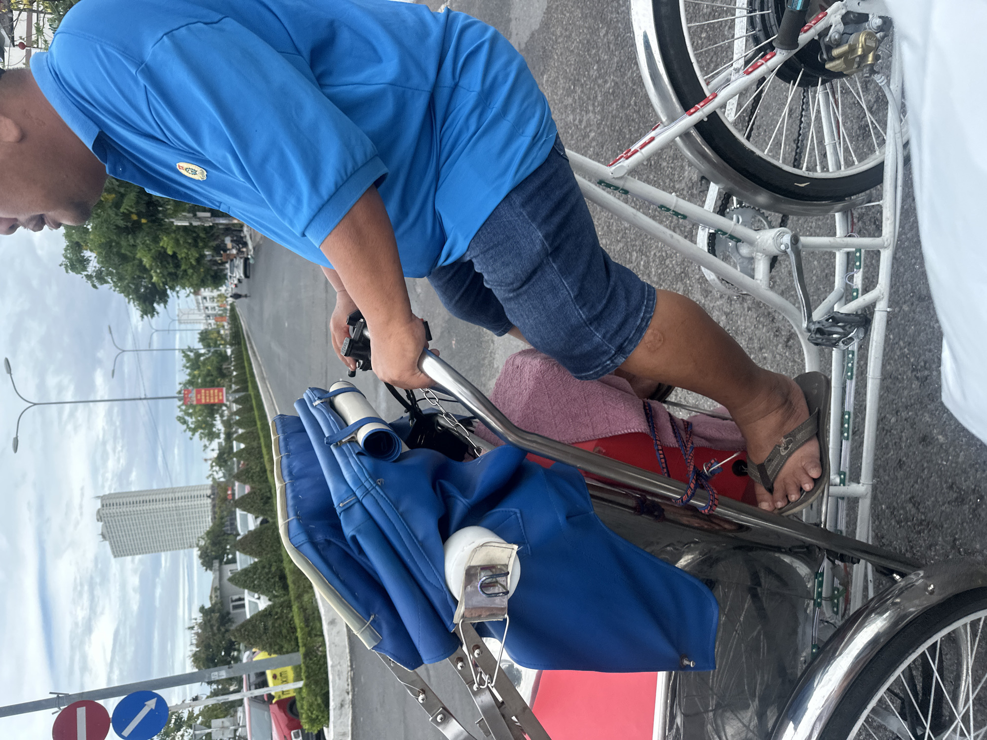 An eye-level shot depicts a man on a pedicab in Nha Trang Ward, Vietnam. The man wears a short-sleeved blue shirt and denim shorts, with brown flip-flops on his feet. He holds the pedicab's handlebar. The pedicab has a white frame, a red passenger seat, and a folded blue canopy. In the background, a street lined with trees and buildings is visible. There are traffic signs on the side of the road, and a tall white building stands in the distance. The sky is partly cloudy.