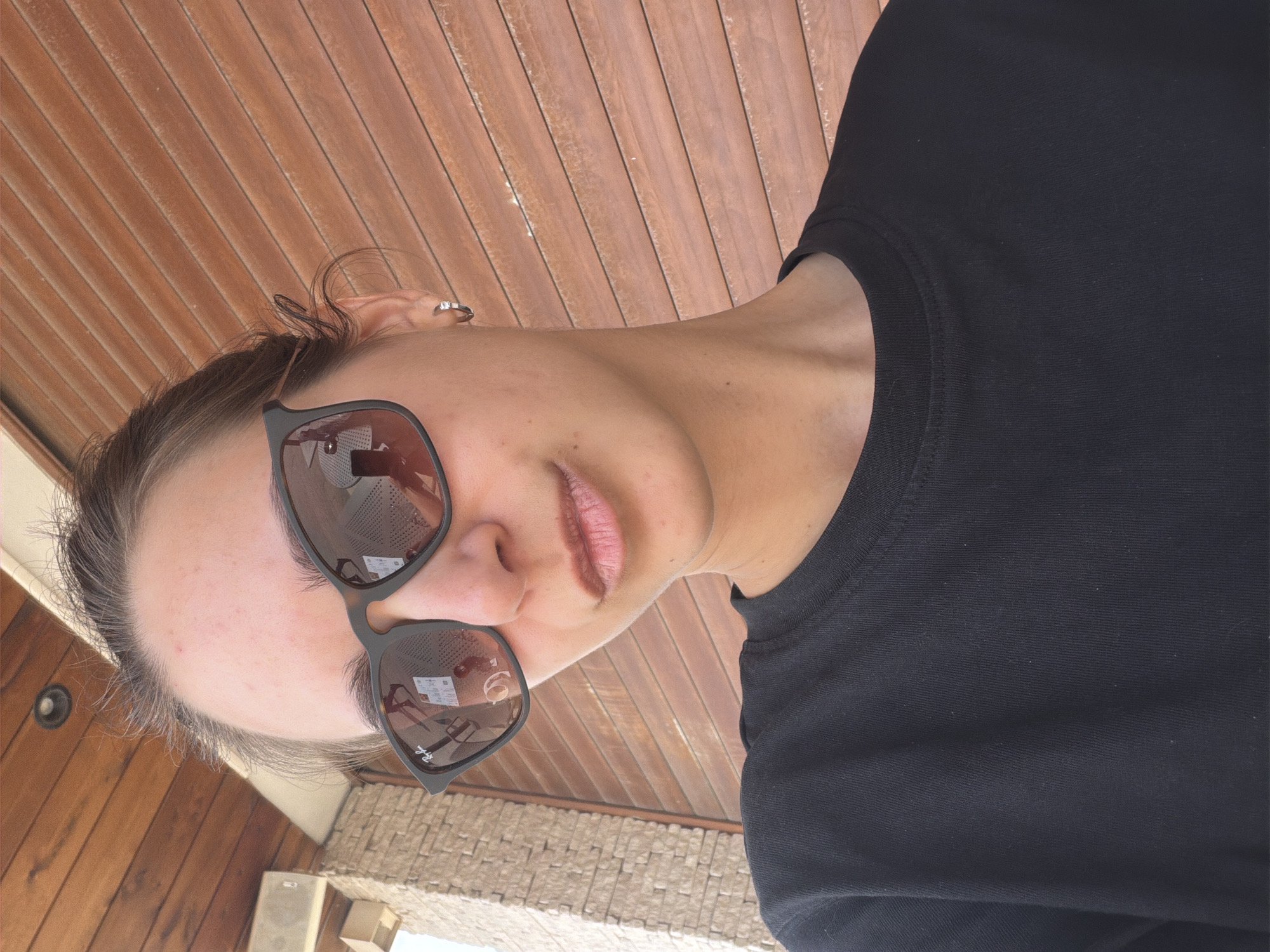 A woman with fair skin and brown hair is depicted from a low-angle perspective. She is wearing sunglasses, which reflect the environment, and a black t-shirt. A silver hoop earring is visible in her left ear. The background consists of wood paneling and, in the lower part of the image, what appears to be a tiled surface and an air conditioning unit, suggesting an outdoor location, possibly Boreti, Montenegro.
