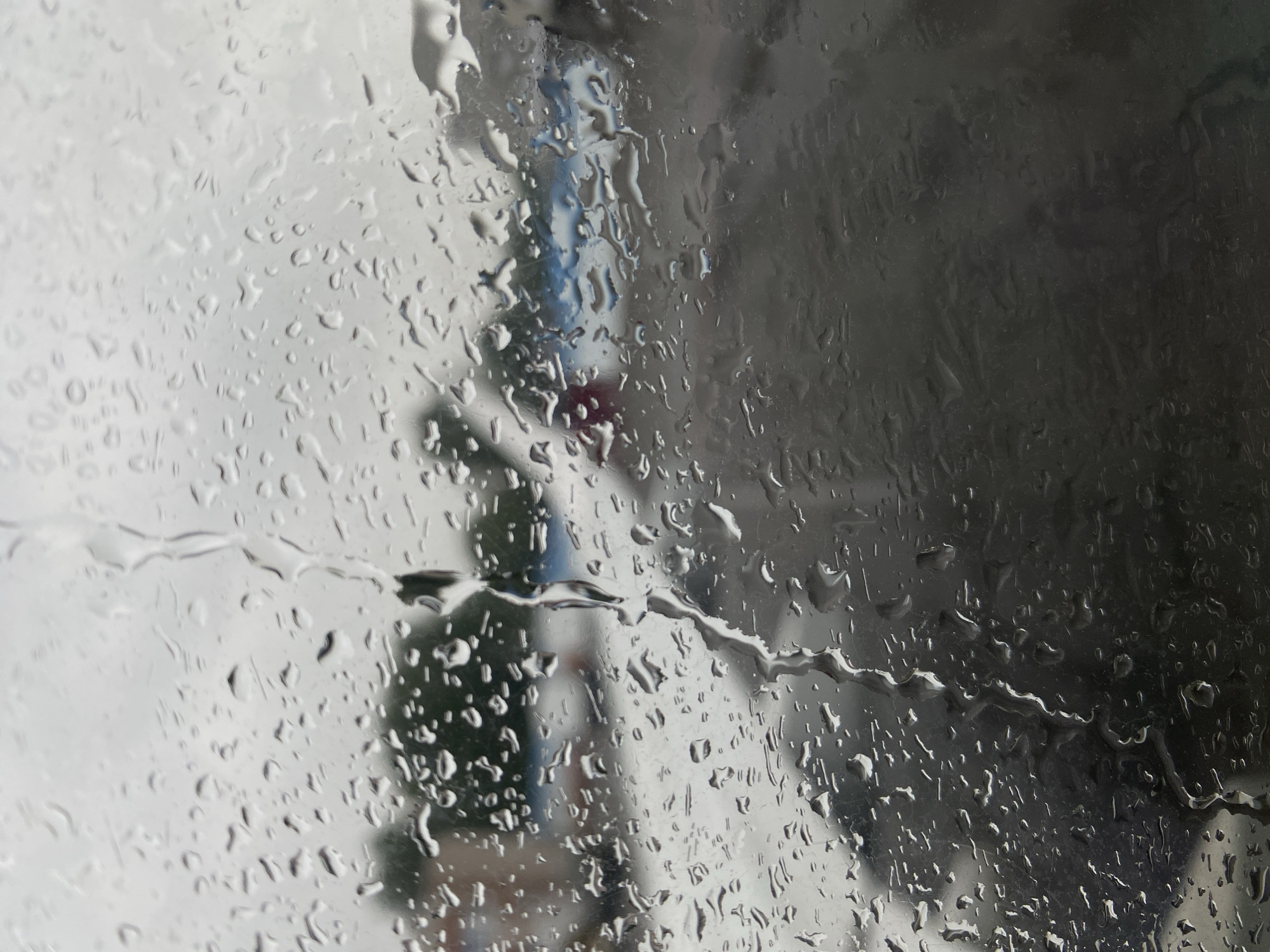 The image is a close-up view of a window covered in raindrops, with a blurred outdoor scene visible through the water droplets. The background suggests an overcast day, possibly in an industrial or semi-rural area. The distant elements include indistinct shapes of buildings, trees, and what appears to be a road or tarmac. There is a hint of a vehicle, possibly an airplane, with a reddish tail fin, in the middle ground. The overall impression is one of a rainy day, obscuring fine details and creating a somewhat melancholic atmosphere. The location is described as Novoye Martyanovo, Russia.