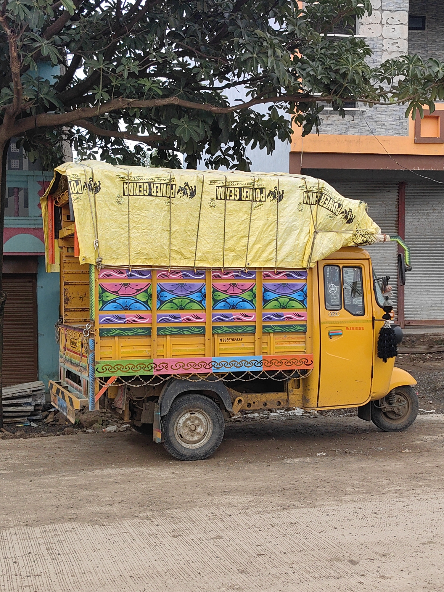 An eye-level, medium shot captures a brightly painted yellow auto-rickshaw truck parked on a dirt road in the city of Padli, India. The truck is adorned with vibrant, colorful patterns on its side panels, featuring motifs of flowers and waves in shades of pink, purple, blue, green, and orange. A large yellow tarp, printed with black text and images of elephants that reads "POWER GENDA," is draped over the truck's cargo area, secured by ropes. The truck has a single driver's cabin with two doors, a side mirror, and a black tassel hanging from the driver's side door. The background shows a building with an orange facade and a grey stone texture, and a tree with large green leaves arches over the truck. The ground is a mix of dirt and a concrete path with horizontal striations. The overall scene depicts a common mode of transportation and commerce in the region.