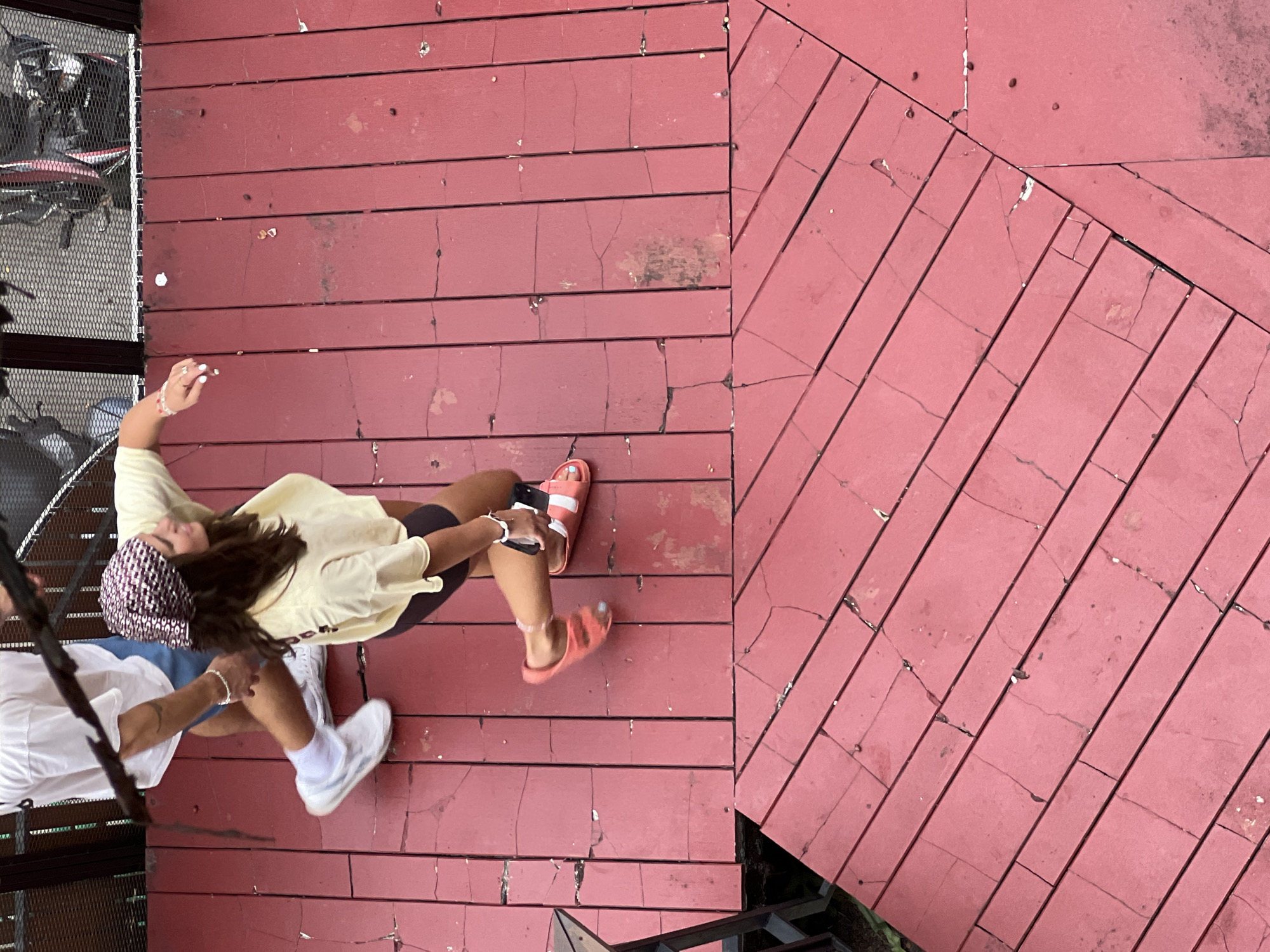 An aerial, high-angle shot shows two people walking on a red wooden deck. The person on the left, wearing a white t-shirt and blue shorts, walks away from the camera with white sneakers and white socks. The person on the right, wearing a yellow t-shirt and dark shorts, walks towards the camera, holding a black phone in their right hand. They wear a patterned bandana on their head and orange slide sandals. The deck is made of planks, some of which are cracked and weathered. In the background, a metal fence with a motorcycle behind it is visible.