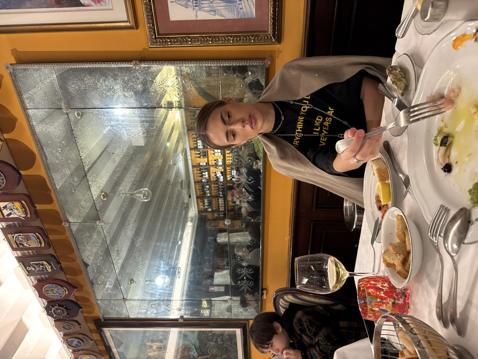 The scene depicts an interior view of a restaurant, identified as Trattoria Da Fiore in Venice, Italy. An adult female, appearing to be in her late 20s to early 30s, is seated at a table on the right side of the frame, actively eating. She has light brown hair pulled back, wears a black long-sleeved shirt with partial text "EVERYTHING YOU LIKED YEARS AGO," and a light brown shawl draped over her shoulders. She wears silver chain necklaces and a ring on her left hand. Her right hand holds a fork with food, poised above a white, partially consumed plate containing pale pink food, a green element, and a dark piece. A knife rests next to her plate.
To her left, a young boy, approximately 5-8 years old, is seated in a striped chair. He has dark hair and wears a brown plaid shirt, with his left hand near his mouth. A stemmed glass containing white wine is positioned between him and the female.
The table, covered with a white tablecloth, holds various dishes and cutlery. Visible items include a silver wire basket with bread rolls, a small white bowl of croutons, a dish with shrimp, lemon wedges, and bread pieces, and a single oyster served in its shell on a white plate. A multi-colored decorative glass stands near the bread basket. A small silver bucket is partially visible between the two diners.
The background features warm yellow-orange walls. Above a wooden wainscoting, a large, multi-paneled antique mirror with etched floral patterns and small decorative rosettes dominates the central wall. Above the mirror, a row of numerous wooden shields or crests with various insignias is affixed to the wall. To the right of the mirror, two framed artworks are visible: the upper one depicts a gondola, and the lower one shows a stylized bell tower, resembling St. Mark's Campanile, indicating a Venetian setting. The mirror reflects the restaurant's interior, including overhead lighting fixtures, a well-stocked bar with numerous bottles, and other indistinct patrons, one of whom appears to be a male behind the bar area.