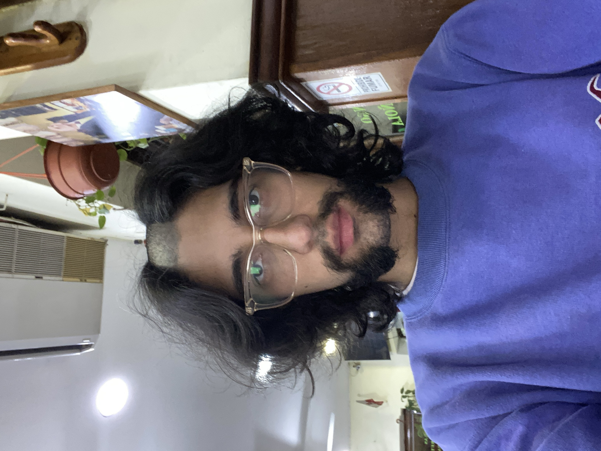 A person with long dark hair, a beard, and glasses is wearing a blue sweatshirt. Behind them, there is a "No Fumar" (No Smoking) sign, a wooden shelf, and a potted plant. An air conditioning unit is visible in the background. The location is likely a building interior in Buenos Aires, Argentina.