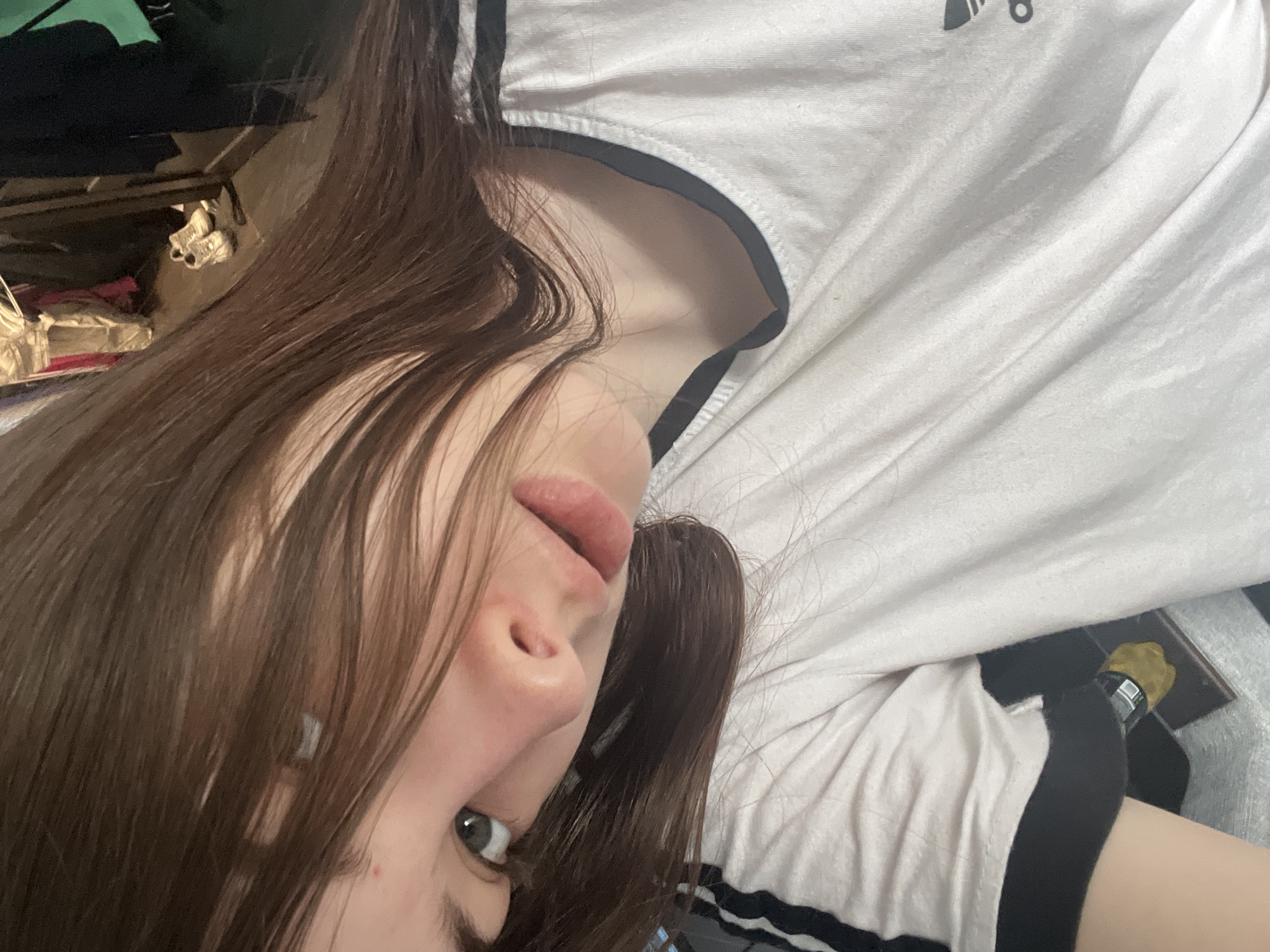 A close-up, angled image features an individual, appearing to be a young female, with long, dark brown hair partially obscuring the left side of her face, including her left eye and forehead. Her visible right eye, with a light iris, is directed towards the camera. Her nose is small, and her lips are slightly parted, revealing a neutral expression. A small, reddish blemish is present on her forehead near her left eyebrow. She is wearing a white, short-sleeved t-shirt with black trim around the collar and black triple stripes on the left sleeve. A partial Adidas logo, black in color, is visible on the lower right area of the shirt. The background, largely out of focus, indicates an indoor environment. To the upper right, various articles of clothing are visible, possibly hanging or draped, and a pair of white athletic shoes rest on a dark floor. A dark, vertical structure, resembling a door frame, is present to the right, with a small glimpse of green behind it. The image contains no visual information to determine the specific geographic location, including the city of Saint Petersburg, Russia.