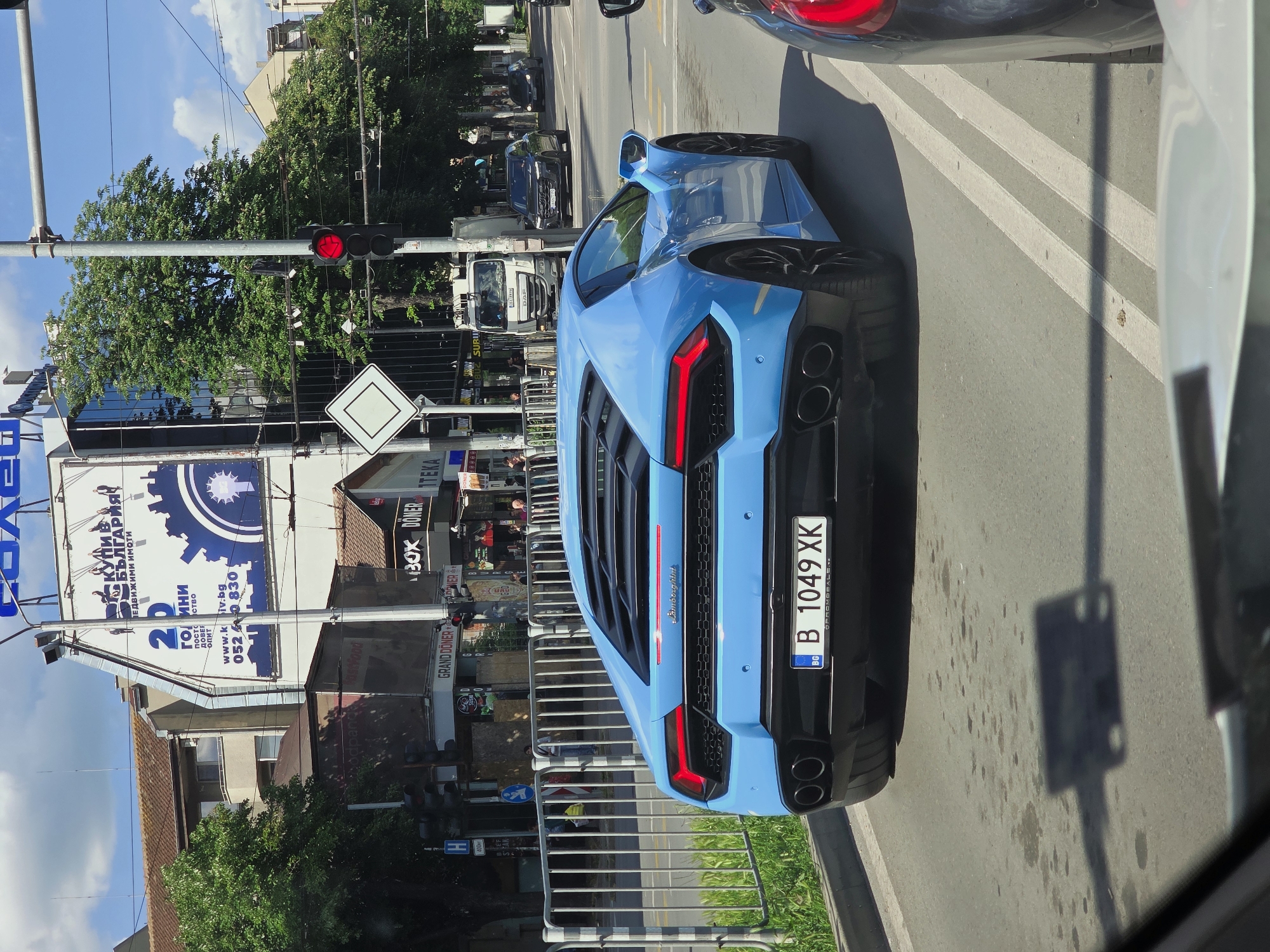 The image shows the rear view of a light blue Lamborghini car on a street in what appears to be an urban setting. The car has a Bulgarian license plate "B 1049 XK." The car is behind a silver car. Behind the Lamborghini, there is a street with other vehicles including a white truck.
On the left of the image, there is a large billboard for a real estate company called "КИВ БЪЛГАРИЯ Недвижими имоти" and a doner restaurant "BOX DONER". A traffic light is showing a red light. There are trees and several buildings in the background. There is also a sign with the text "GRAND DONER".
Based on the signs and license plate, the location is in Bulgaria, likely in the city of Varna given the phone code 052 on the real estate billboard. The presence of a doner restaurant also suggests a central urban area.
