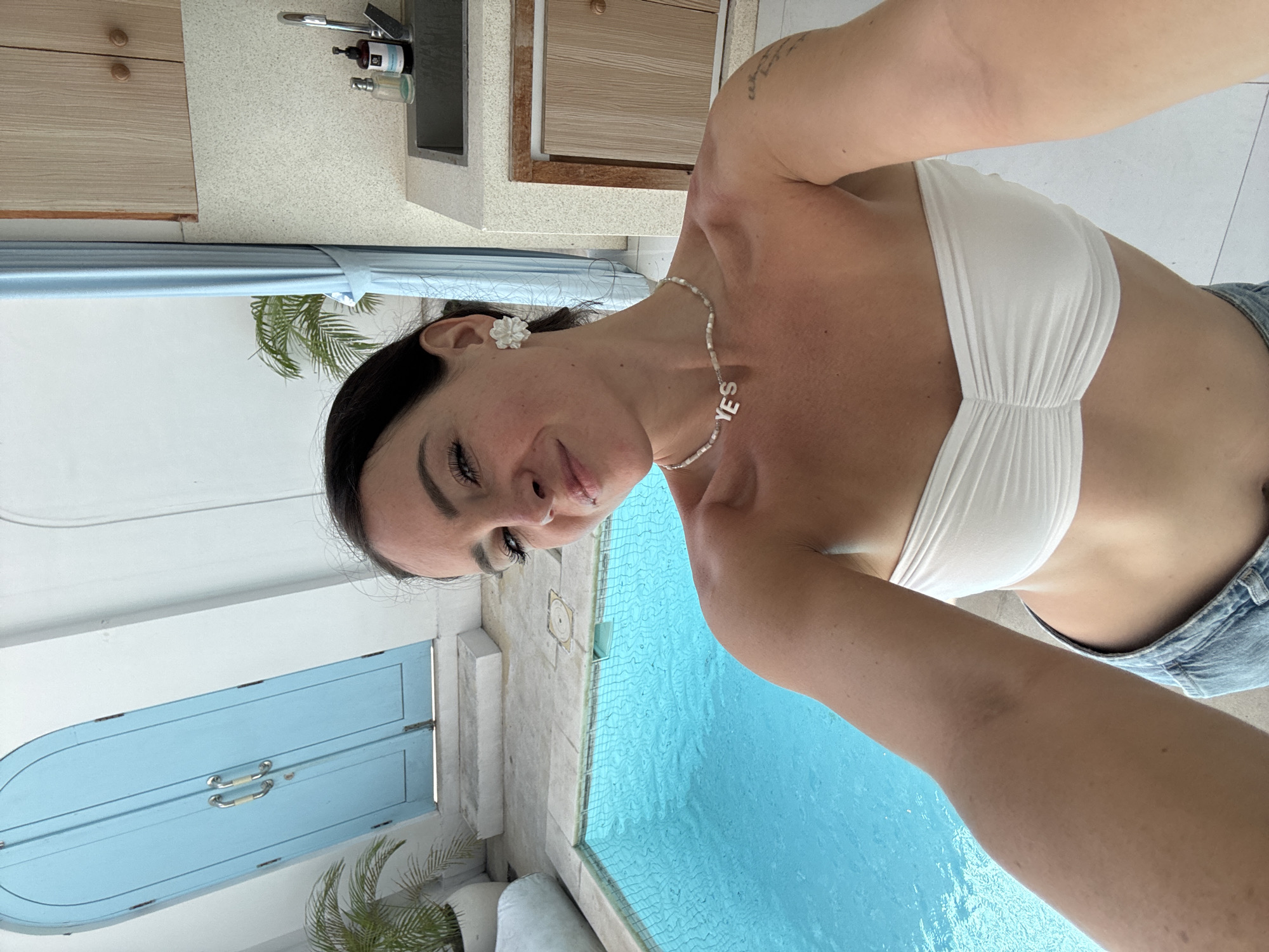 A woman with dark hair is taking a selfie in an outdoor setting. She is wearing a white strapless top and blue denim shorts. Around her neck, she has a pearl necklace with "YES" spelled out in white block letters. She also has a white flower-shaped earring in her left ear. The background shows a bright blue swimming pool, a light blue arched door, and some plants. To the right, there is a sink with a faucet and wooden cabinets. The overall impression is one of a relaxed and sunny environment, possibly a resort or a private villa. The image appears to be taken in Mengwi, Indonesia.