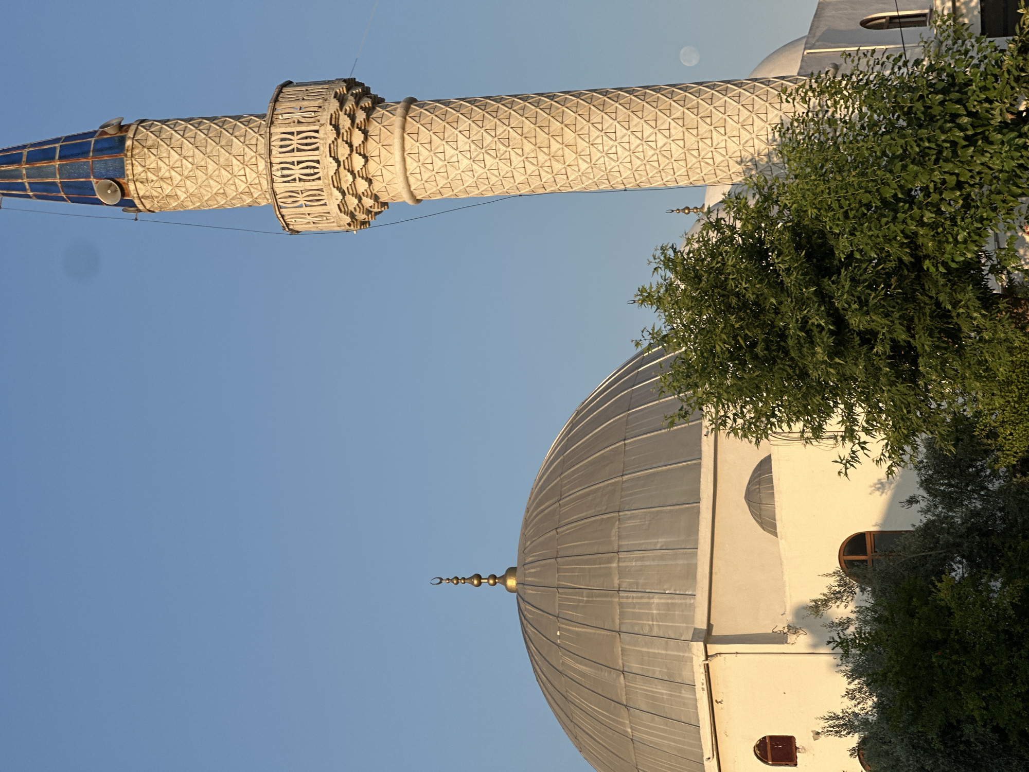 A medium shot captures the exterior of a mosque in Fethiye, Turkey, bathed in the warm, low light of either sunrise or sunset. The image is dominated by the mosque's architecture, which includes a large, silver-colored dome topped with a crescent moon finial and a tall, intricately patterned minaret with a blue-capped spire. The minaret features a balcony with decorative railings.
In the foreground, lush green trees partially obscure the lower portion of the mosque's white facade. The mosque itself has arched windows and what appears to be another smaller dome visible behind the trees. The sky is a clear, serene blue. A faint full moon is visible in the upper right portion of the frame. The overall impression is one of tranquil beauty and architectural splendor.