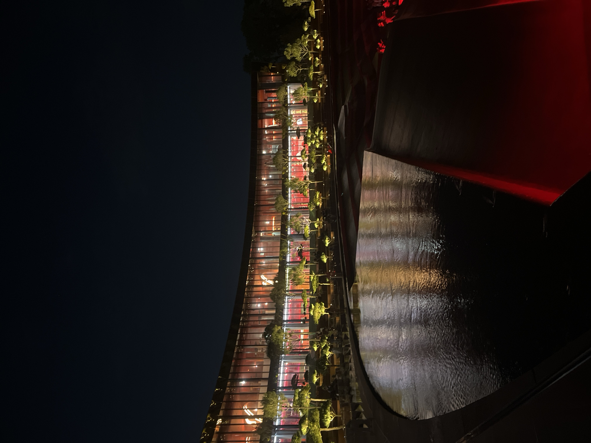 This is a nighttime, wide-angle, exterior photograph of a modern, curved building with a glass facade. The building is illuminated from within with red and warm white lights, creating reflections on the surface of a dark, reflective pool of water in the foreground. The pool's edge is lined with what appear to be tiered planters or seating areas, populated with small, ornamental trees that are individually lit with warm yellow lights. The landscaping suggests a manicured, possibly formal, garden setting. The building itself has multiple stories visible through the glass, with internal lighting highlighting structural elements and interior spaces. The sky above is dark and clear, indicating nighttime. The overall aesthetic is one of contemporary architecture with dramatic lighting, possibly a restaurant, entertainment venue, or a public building designed for nighttime viewing. The location is the Krasnodar Expo exhibition center in Krasnodar, Russia.
