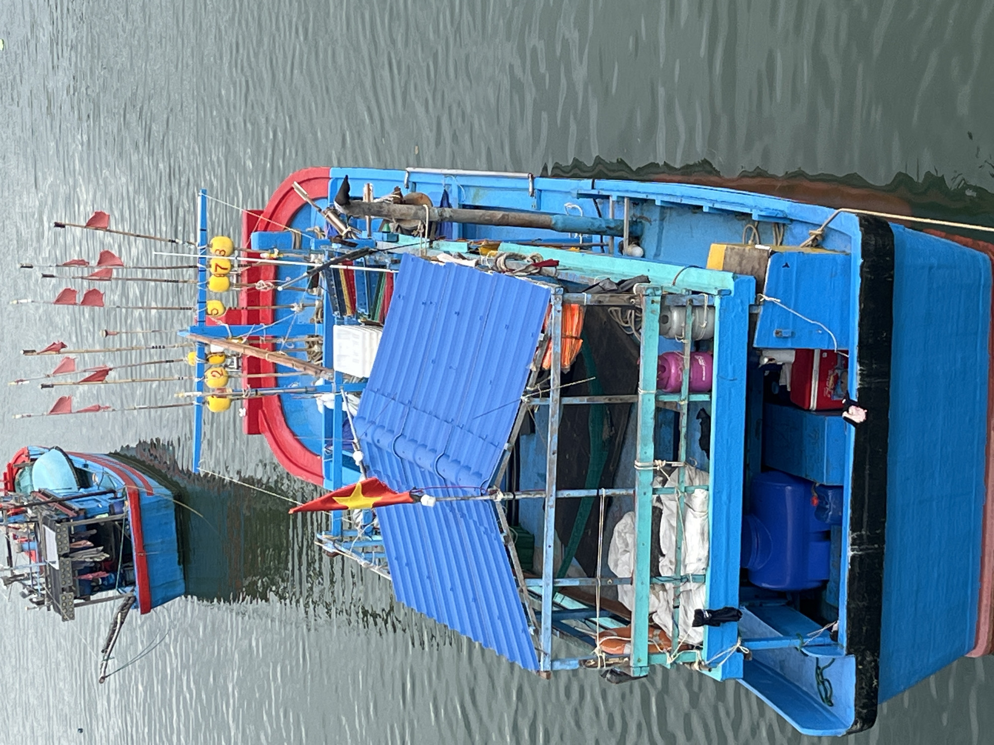 This is a high-angle, close-up shot of two blue and red fishing boats docked in calm, grey water, likely in the city of Nha Trang, Vietnam. The boat in the foreground is larger and more detailed. It has a blue, corrugated metal roof covering a central structure. A Vietnamese flag flies from a pole on top of this roof. The boat is equipped with various fishing gear, including numerous poles extending from the stern, some with red flags attached, and yellow buoys. There are life preservers, storage containers, and other equipment visible on deck. The boat has a bright blue hull with a red trim line near the waterline. The boat in the background is smaller and appears to be in the process of being repaired or loaded, with some of its internal structure visible. The water is slightly choppy, creating ripples around the boats. There are no people visible in the image.