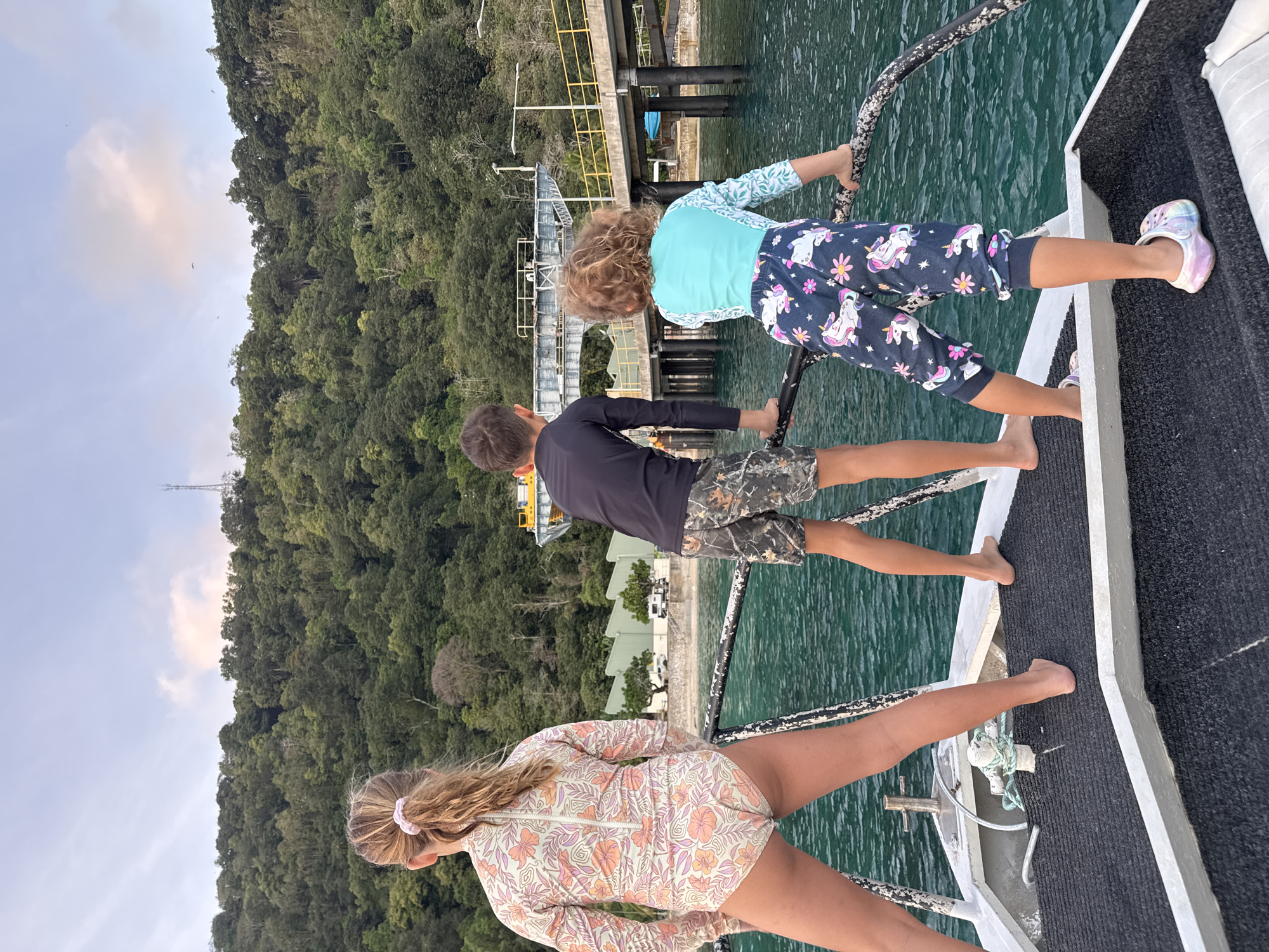 Three children are standing on the deck of a boat, looking out at a forested hillside and a pier with industrial structures. The child on the left is a girl facing away from the camera, wearing a long-sleeved swimsuit with a floral pattern. The child in the middle is a boy facing away from the camera, wearing a dark long-sleeved shirt and camouflage shorts. The child on the right is a girl facing away from the camera, wearing a light blue long-sleeved shirt and dark patterned pants with unicorns and flowers. The water is a clear turquoise. The hillside is covered in dense green vegetation. A pier with yellow railings and some yellow industrial equipment extends into the water. The sky is partly cloudy. This scene is likely on Christmas Island, an Australian territory.