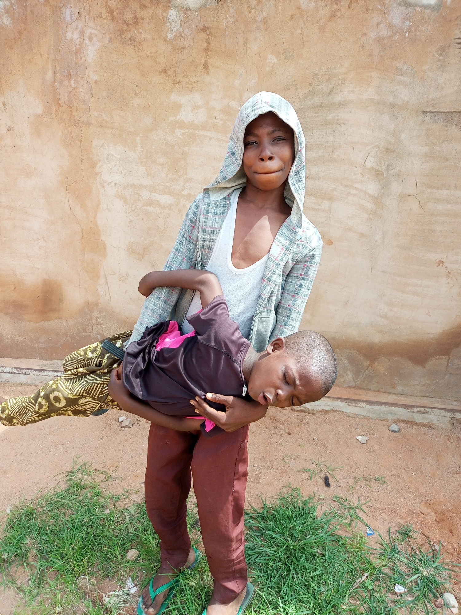 A young child, appearing to be a boy, is being carried by an older child, who is wearing a hooded jacket over a white tank top and patterned pants. The older child is looking directly at the camera with a slight smile. The younger child, who is asleep or unconscious, is wearing a dark t-shirt with a pink accent and patterned trousers. They are standing on a dirt and grass patch, with a textured, light brown wall behind them. The location is specified as the city of Gombe, Nigeria.