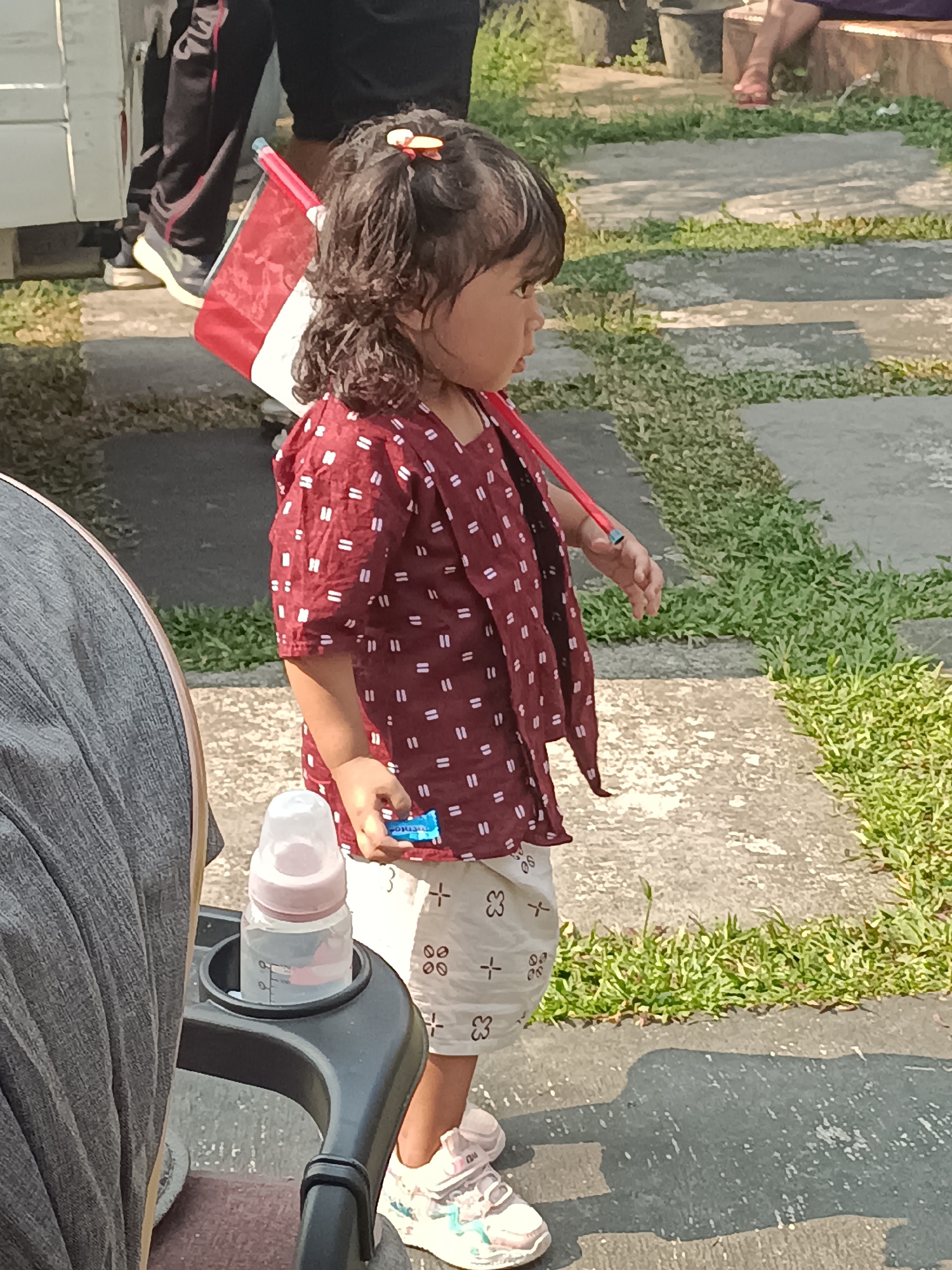 A young girl stands on a paved path outdoors, holding a small Indonesian flag. She has dark, curly hair tied back with a red clip and is wearing a maroon shirt with a white pattern and white shorts with a similar pattern. She is looking to her right, with a neutral expression. A stroller is partially visible on the left, with a baby bottle in its holder. In the background, people are seen standing, and there are potted plants. The scene appears to be in a garden or park setting, with grass and stone paving. The lighting suggests it is daytime, likely sunny. The location is identified as Sleman, Indonesia. There is no discernible text in the image.
