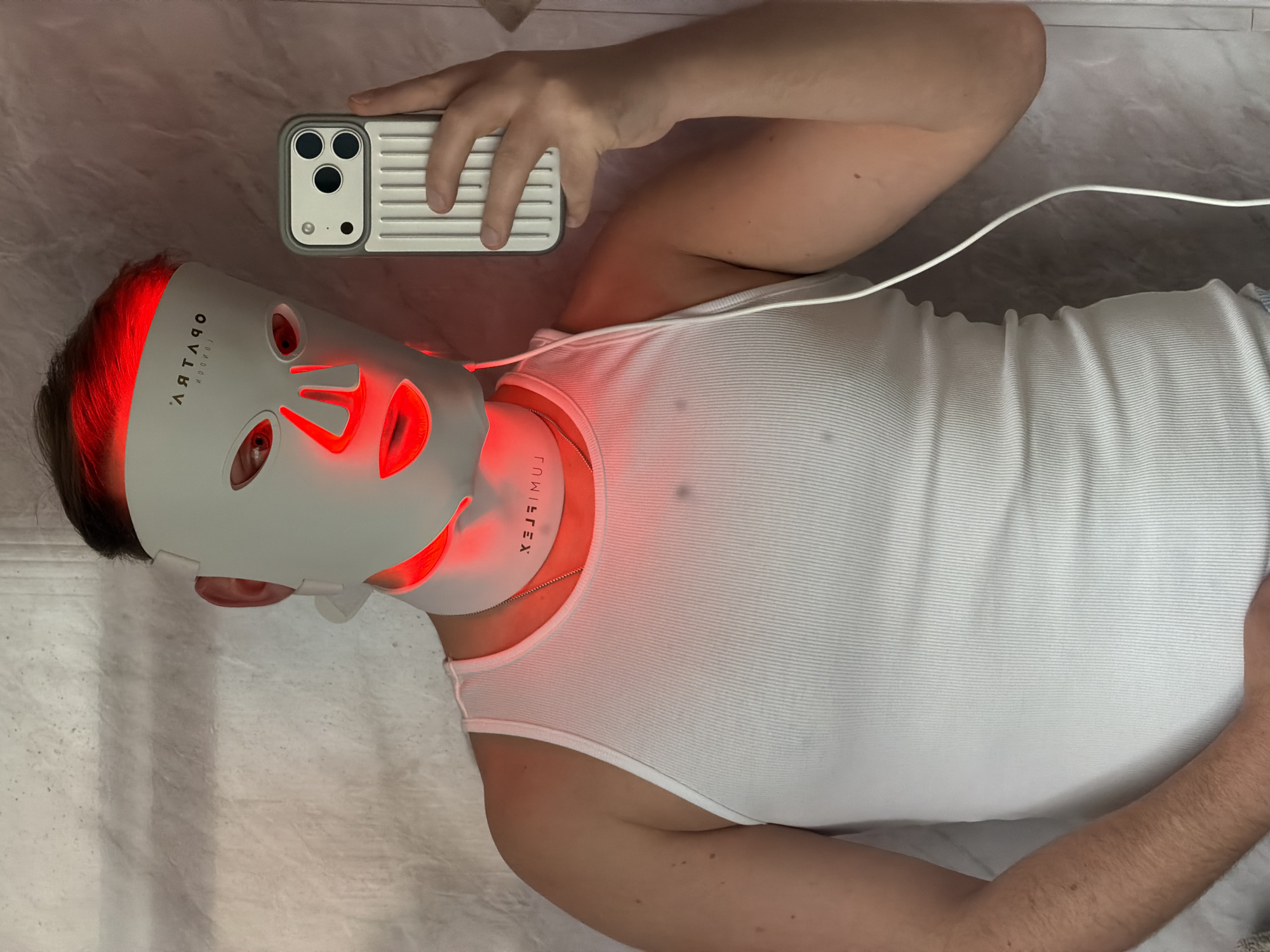 A person wearing a white tank top and an illuminated red light therapy mask lies on a white surface, possibly a bed. The mask covers the person's face, with red lights emitting from the forehead, eyes, nose, and mouth areas. The mask has text on it, including "ARTARQ" and "LUMIFLEX." A white cable connects to the mask. The person holds a white and gray phone case with multiple camera lenses above their head, possibly taking a selfie. The background appears to be a neutral, textured surface. The image does not contain sufficient information to identify a specific location in London.