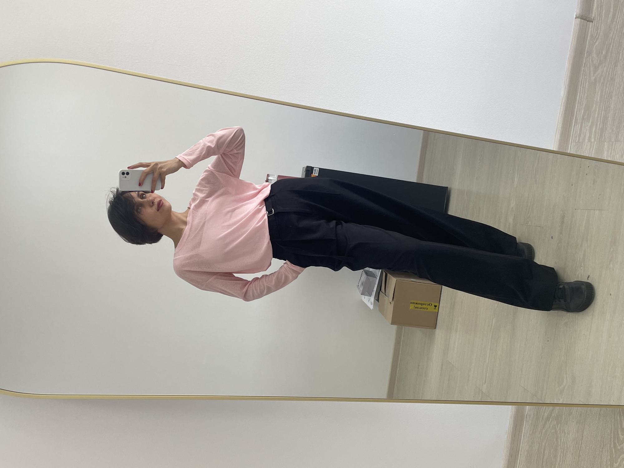 A person with short, dark hair is taking a selfie in a mirror. The person is wearing a long-sleeved, light pink shirt and black pants. The person is holding a white smartphone in their right hand, positioning it to capture their reflection. The mirror has a thin, gold frame. The background shows a white wall and a light-colored floor, possibly made of wood or laminate. Several boxes and other items are visible near the bottom of the mirror's reflection. The location is unknown and there is no evidence to confirm it is in Izhevsk, Russia.